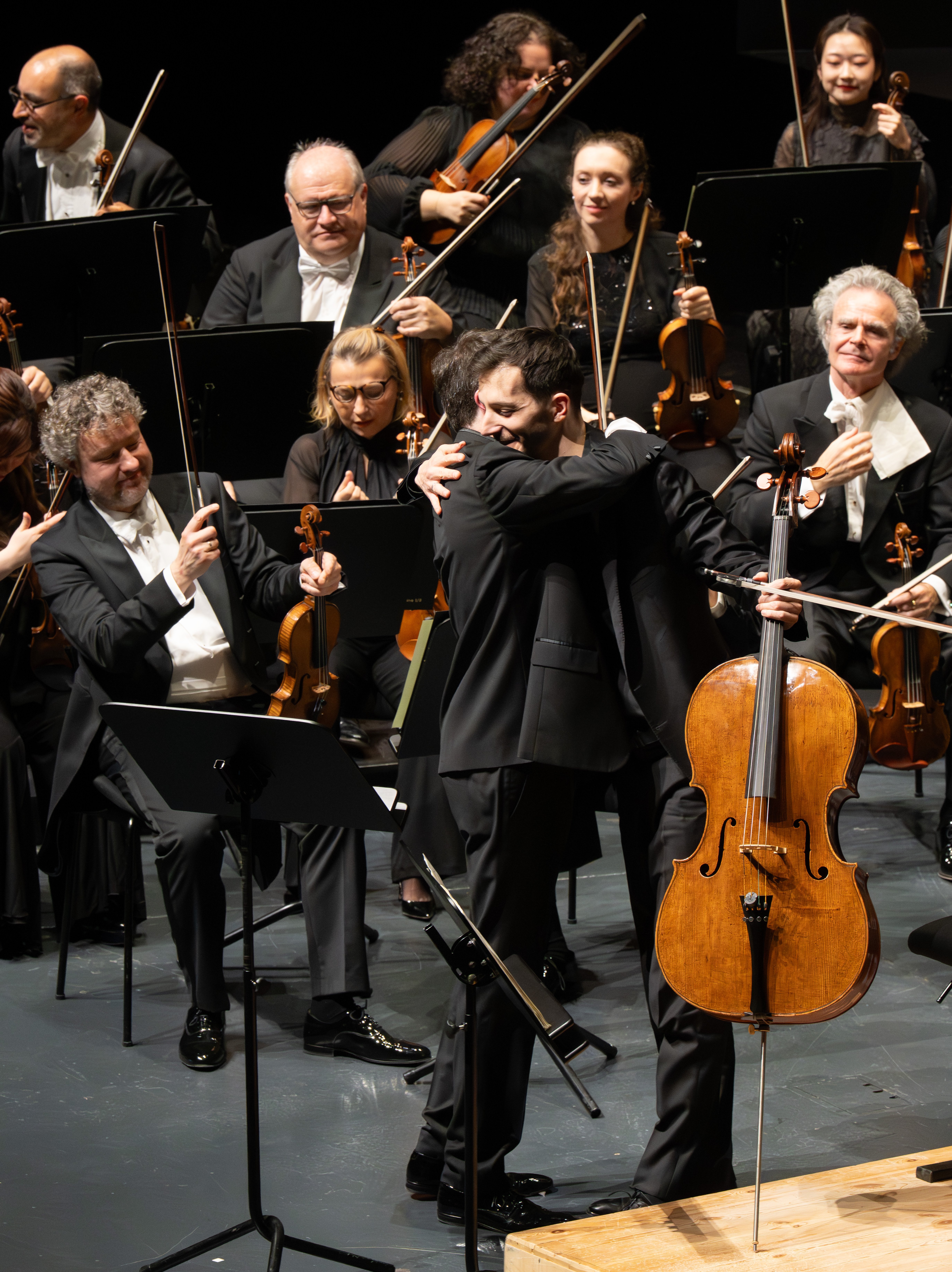Zwei Musiker umarmen sich auf der Bühne, einer von ihnen hält ein Cello. Um sie herum stehen Orchestermitglieder mit Streichinstrumenten.