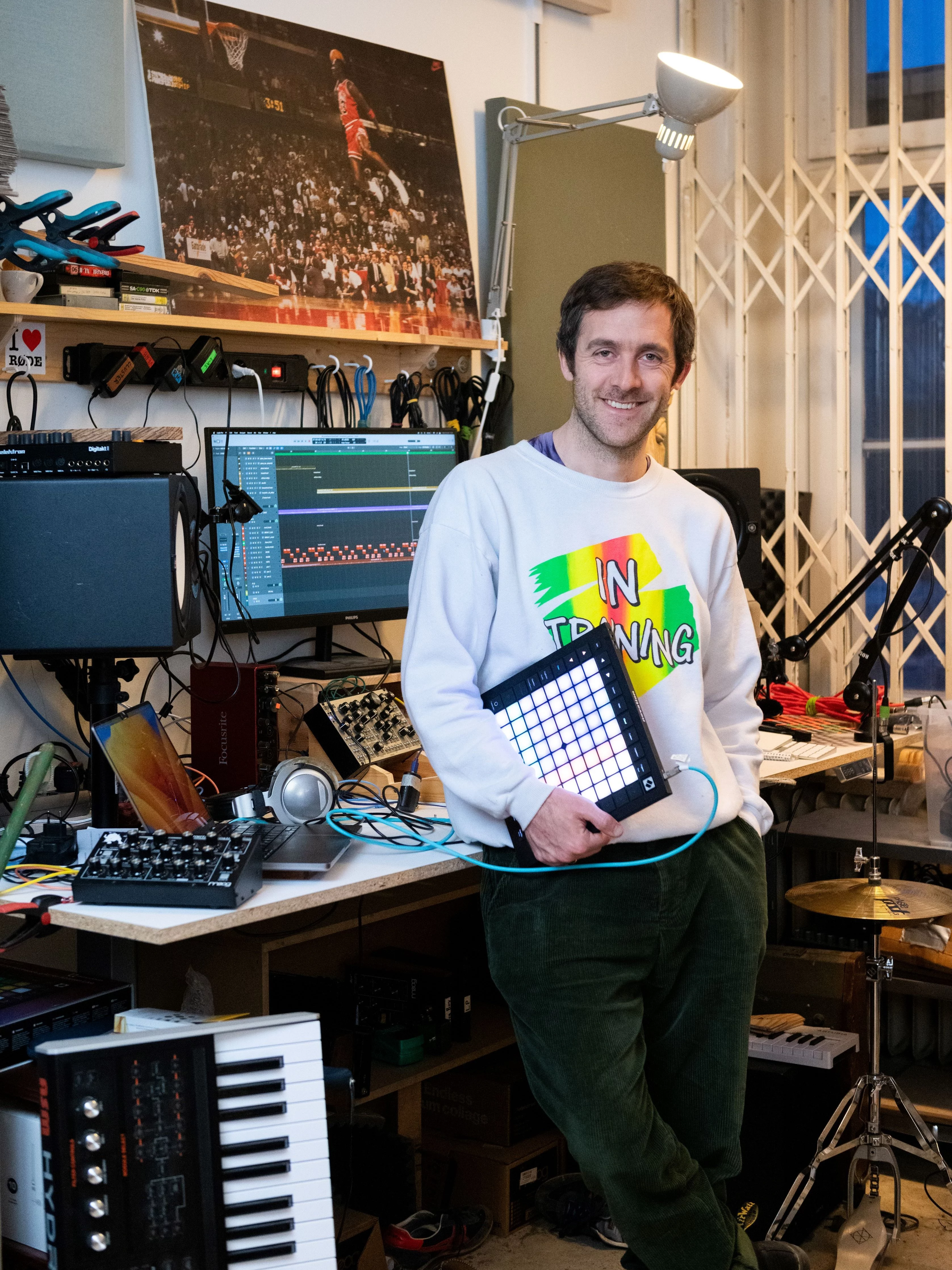 Allan Nilles steht in einem Musikstudio, hält einen Pad-Controller und lächelt. Im Raum befinden sich verschiedene Audiogeräte, ein Computer, Keyboards, ein Schlagzeug und ein Basketball-Poster an der Wand.