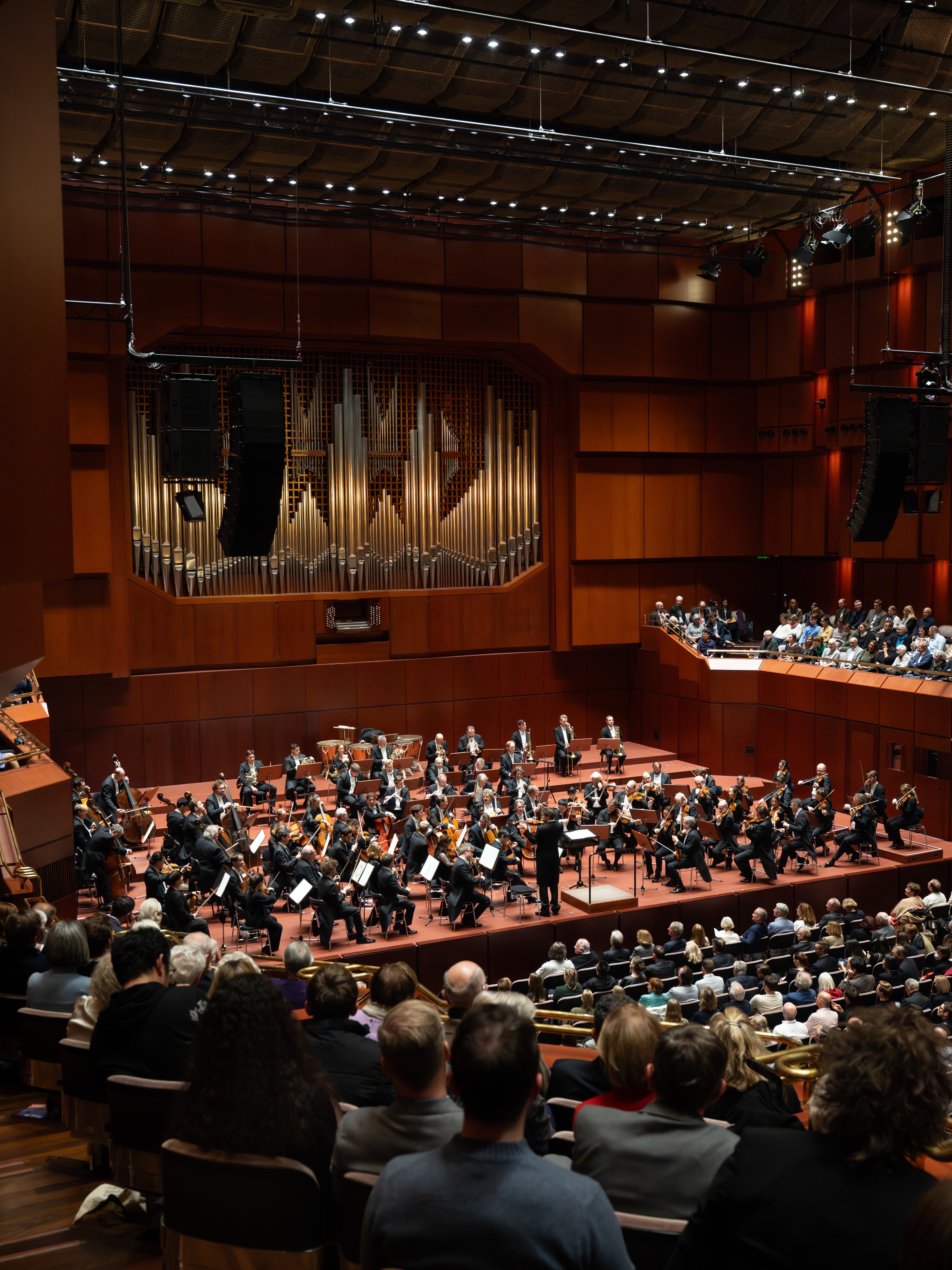The Berliner Philharmoniker performs on stage in a large concert hall with seated audience members. The stage features a grand pipe organ and musicians arranged in sections under warm lighting.