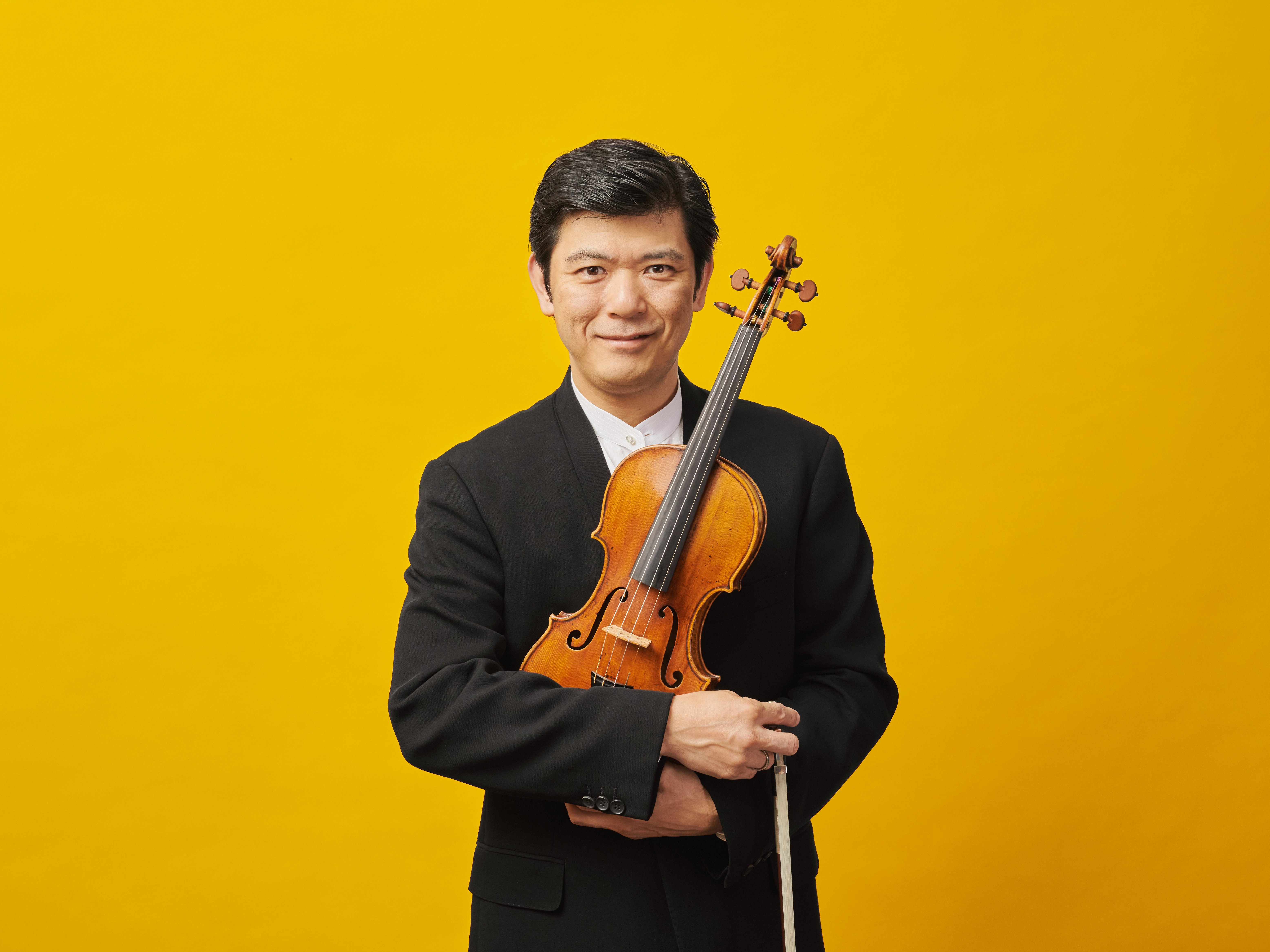 Daishin Kashimoto, mit schwarzen Haaren und im Frack, hält seine Geige in der Hand und lächelt in der Kamera. Er steht von einem leuchtend gelben Hintergrund.