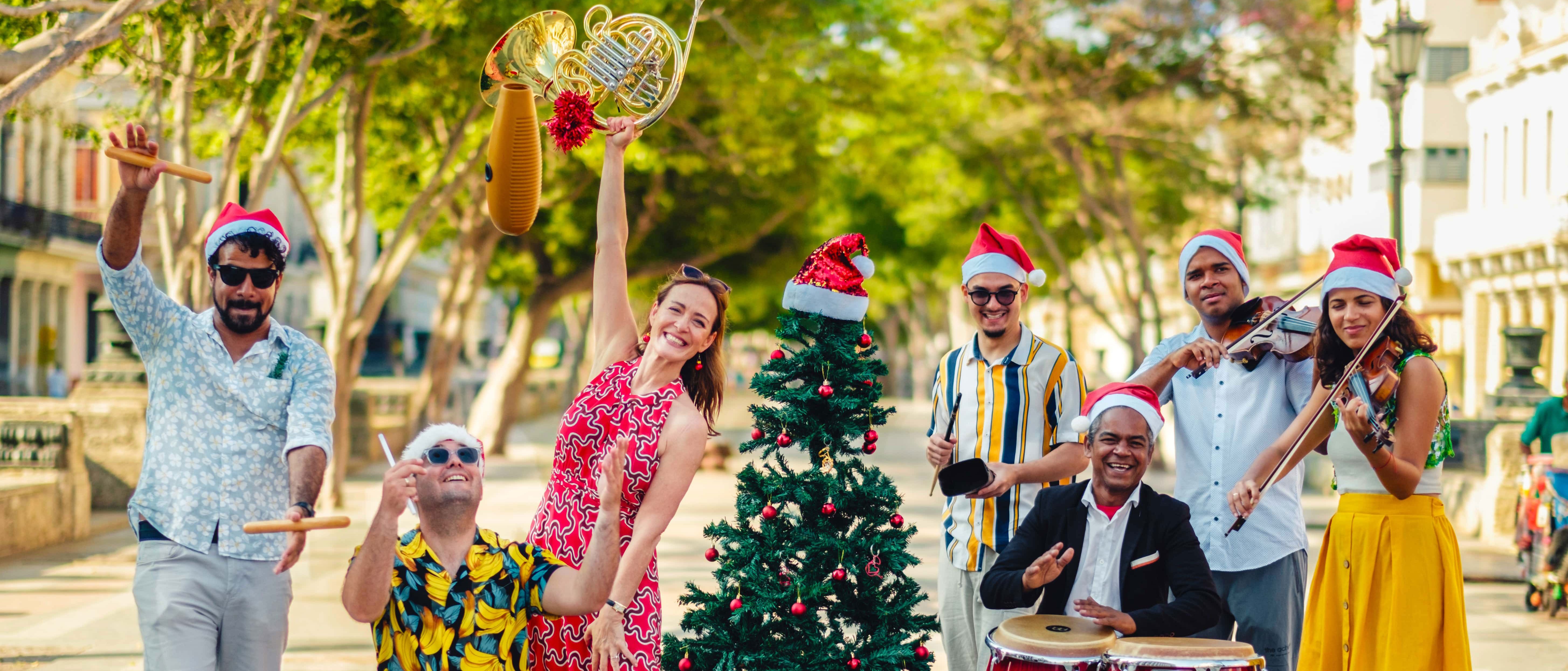 A cheerful group of seven people in festive clothes and Santa hats play musical instruments around a decorated Christmas tree outdoors on a sunny day.