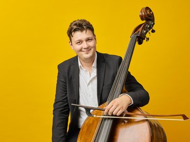 Ein Mann in einem schwarzen Anzug und weißem Hemd lächelt, während er einen Kontrabass vor einem leuchtend gelben Hintergrund hält und spielt.