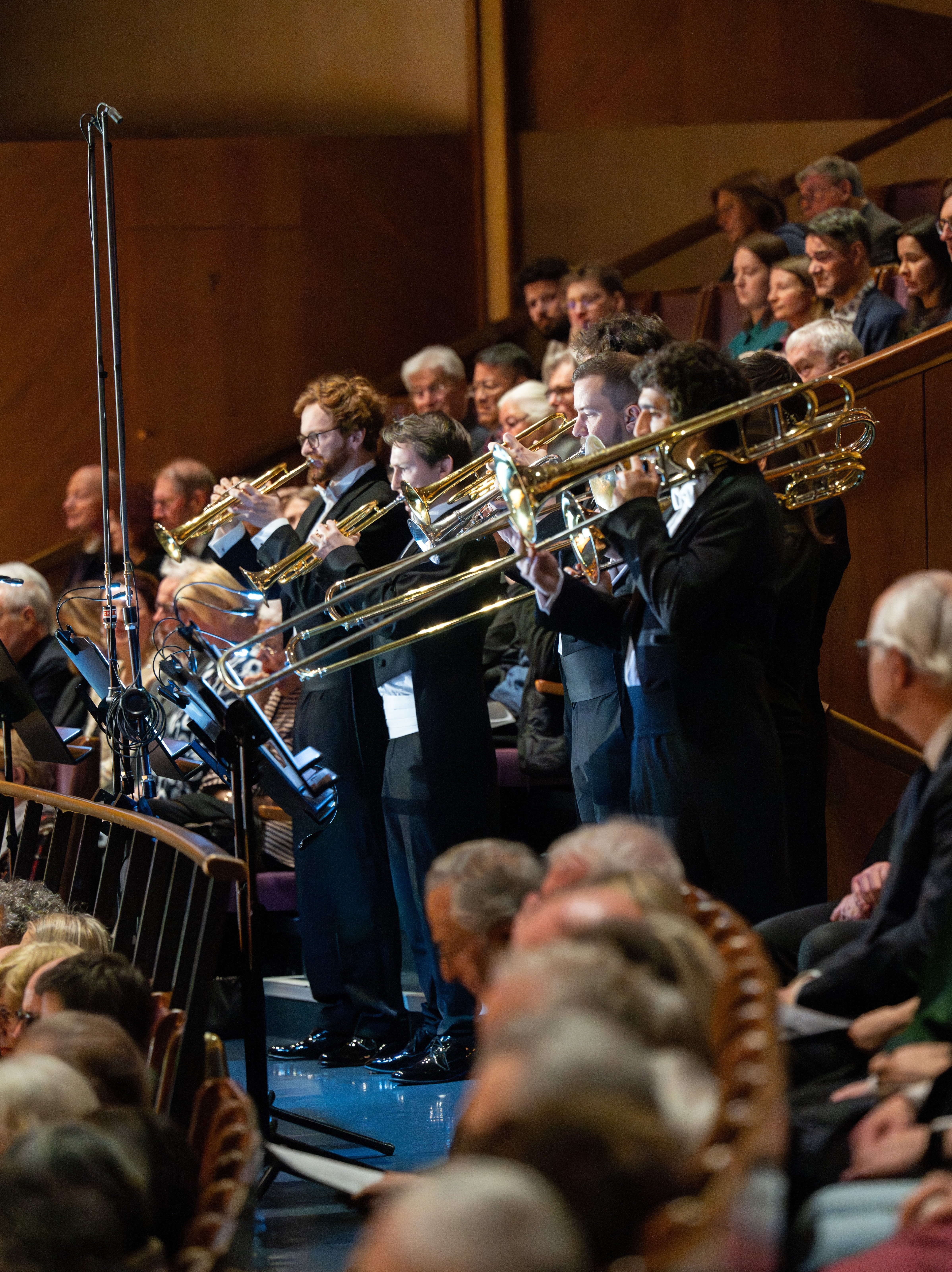 Posaunenbläser in förmlicher Kleidung spielen in einem Orchester, inmitten eines sitzenden Publikums in einem holzgetäfelten Konzertsaal mit Zuschauerreihen.