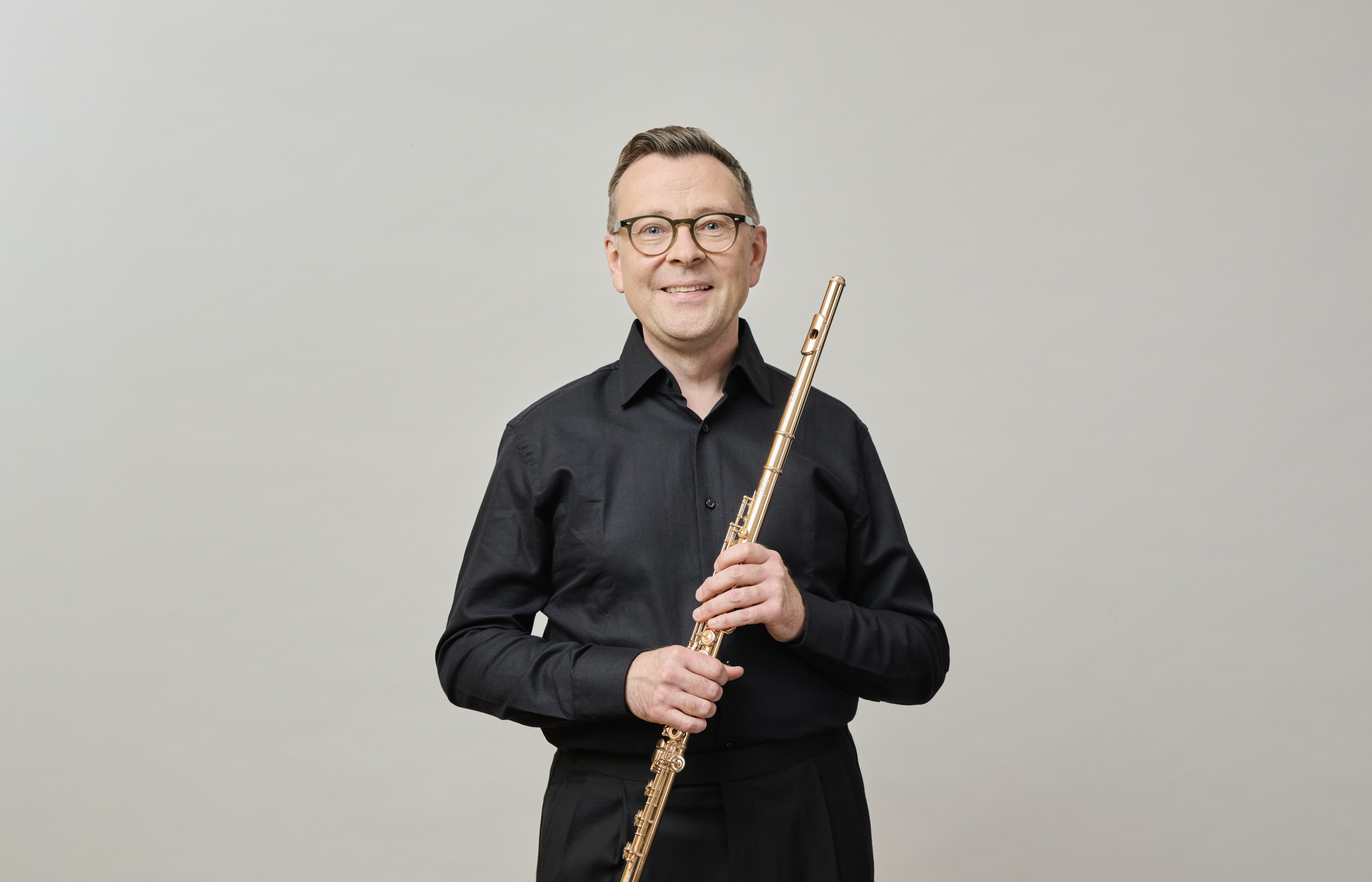 Ein Mann mit Brille und schwarzem Hemd steht vor einem schlichten Hintergrund, lächelt und hält eine Flöte senkrecht in beiden Händen.