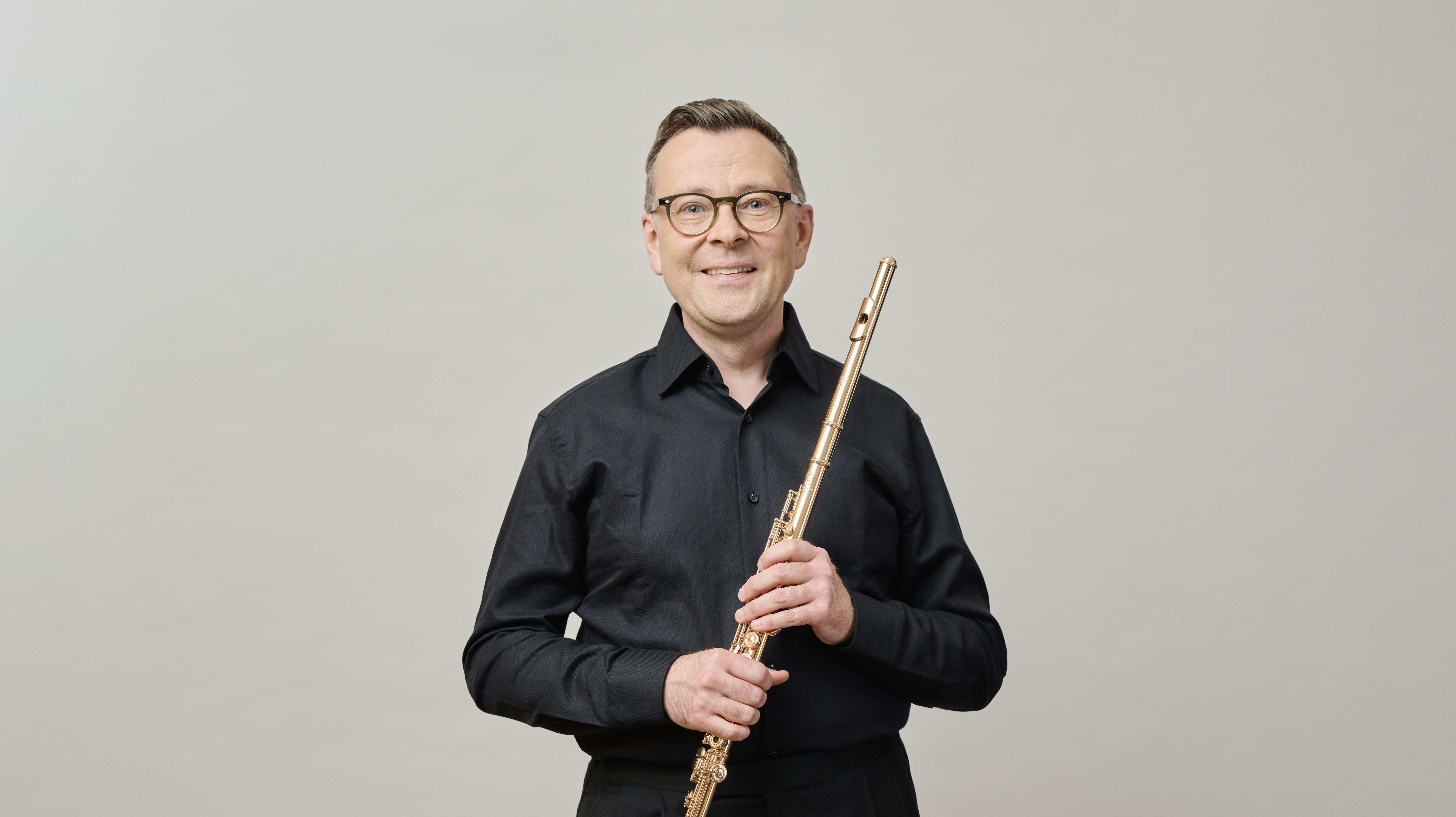 Ein Mann mit Brille und schwarzem Hemd steht vor einem schlichten Hintergrund, lächelt und hält eine Flöte senkrecht in beiden Händen.
