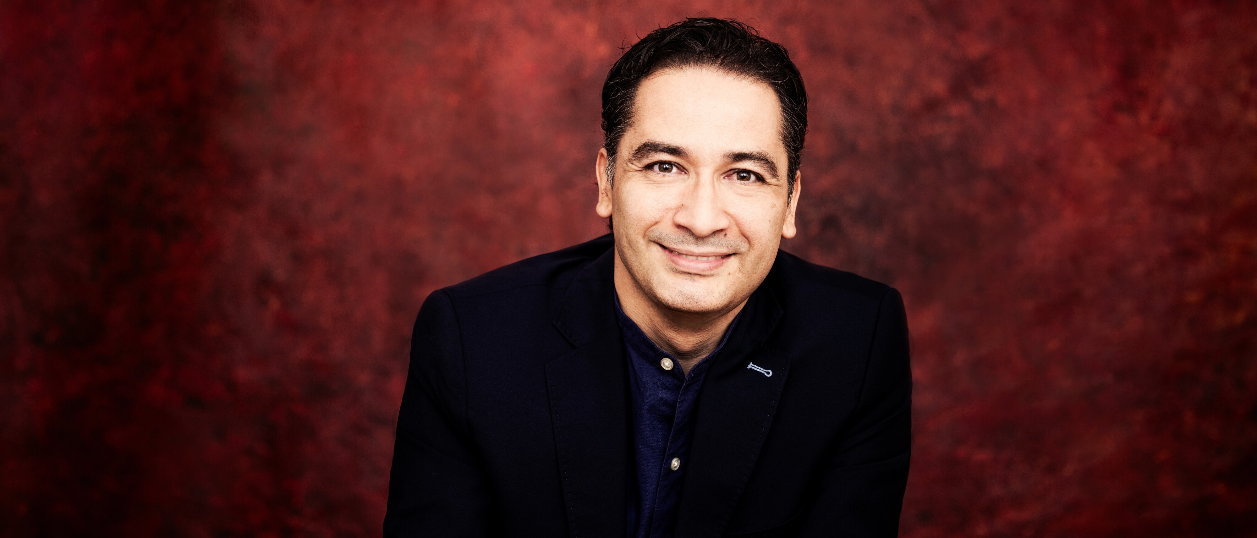 Andrés Orozco-Estrada mit dunklem Haar, der einen dunklen Anzug und ein dunkles Hemd trägt, sitzt mit verschränkten Händen da und lächelt in die Kamera vor einem tiefroten, strukturierten Hintergrund.