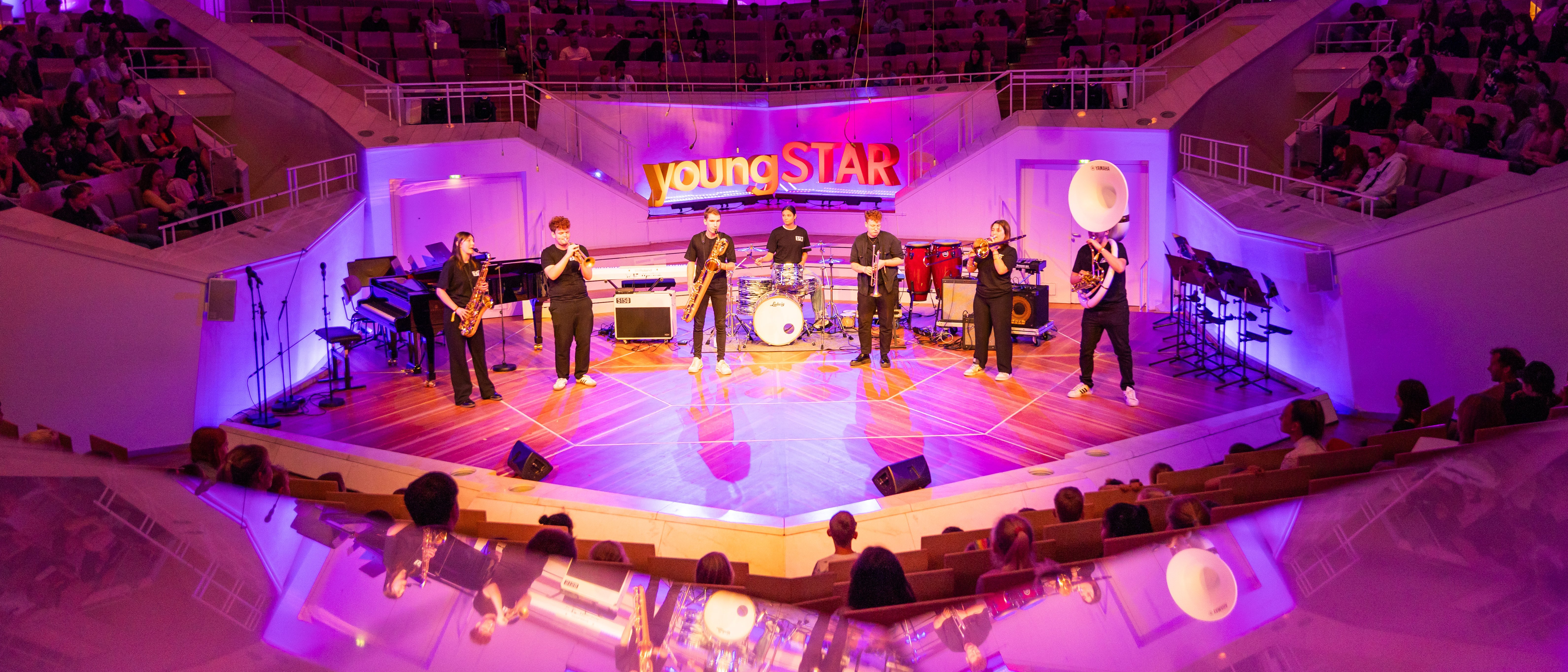 A band performs on a brightly lit stage with purple lighting in a modern concert hall before an audience. Musical instruments, including drums and guitars, are visible on stage.
