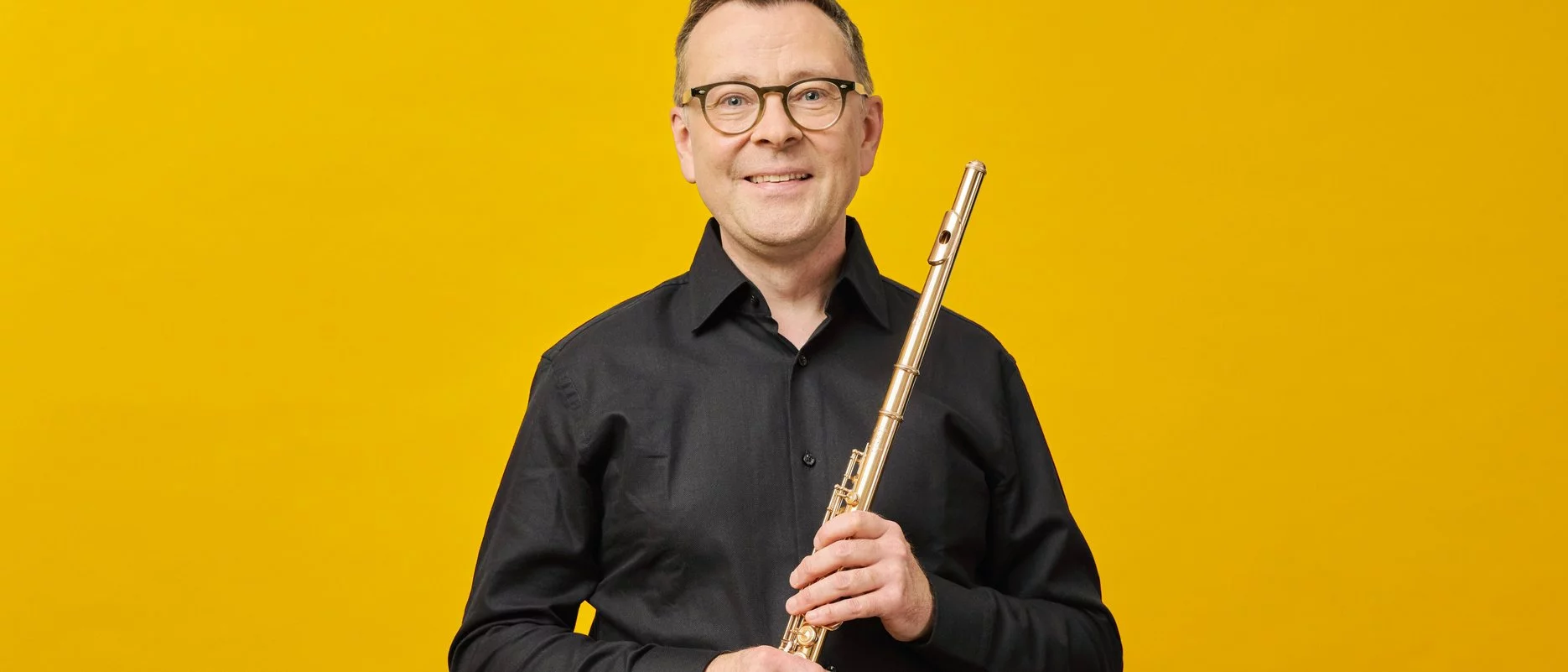 A smiling Stefán Ragnar Höskuldsson in glasses and a black shirt holds a flute upright in front of a bright yellow background.