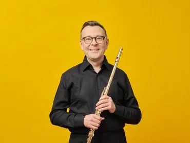 A smiling Stefán Ragnar Höskuldsson in glasses and a black shirt holds a flute upright in front of a bright yellow background.