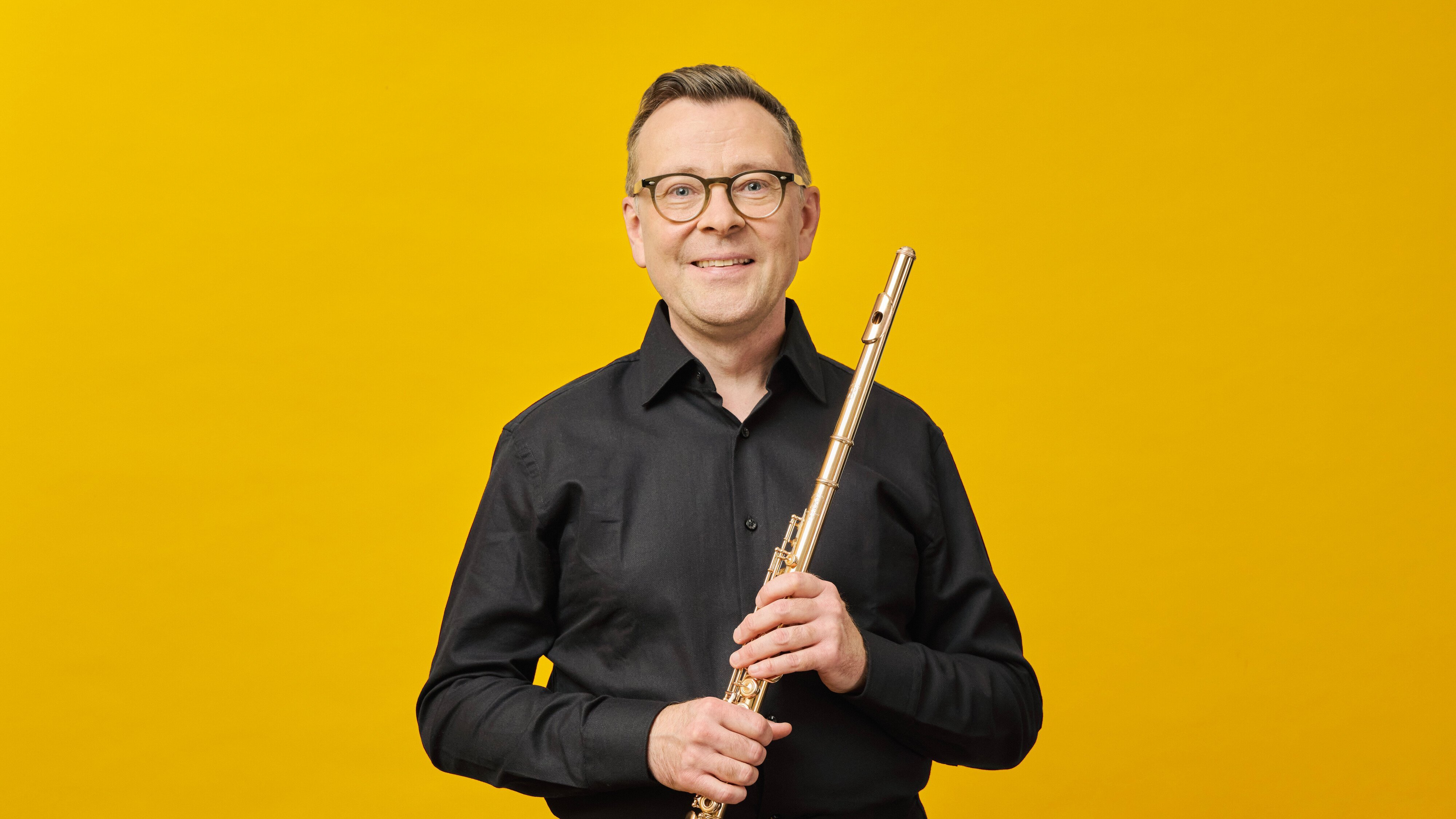 Stefán Ragnar Höskuldsson mit Brille und schwarzem Hemd hält eine Flöte vor einem leuchtend gelben Hintergrund.