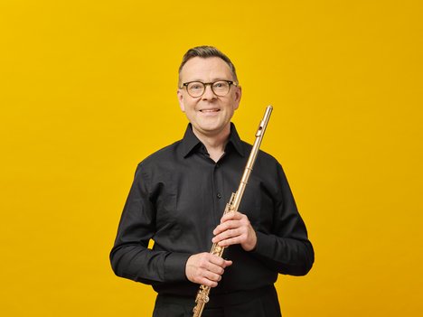 A smiling Stefán Ragnar Höskuldsson in glasses and a black shirt holds a flute upright in front of a bright yellow background.