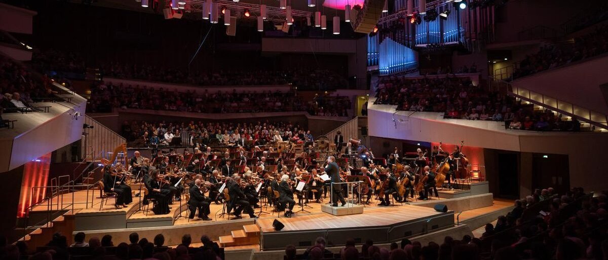 Ein vollbesetztes Orchester spielt auf der Bühne eines großen, modernen Konzertsaals, umgeben von Zuschauern, unter dramatischer Beleuchtung und unter hängenden Deckenpaneelen.