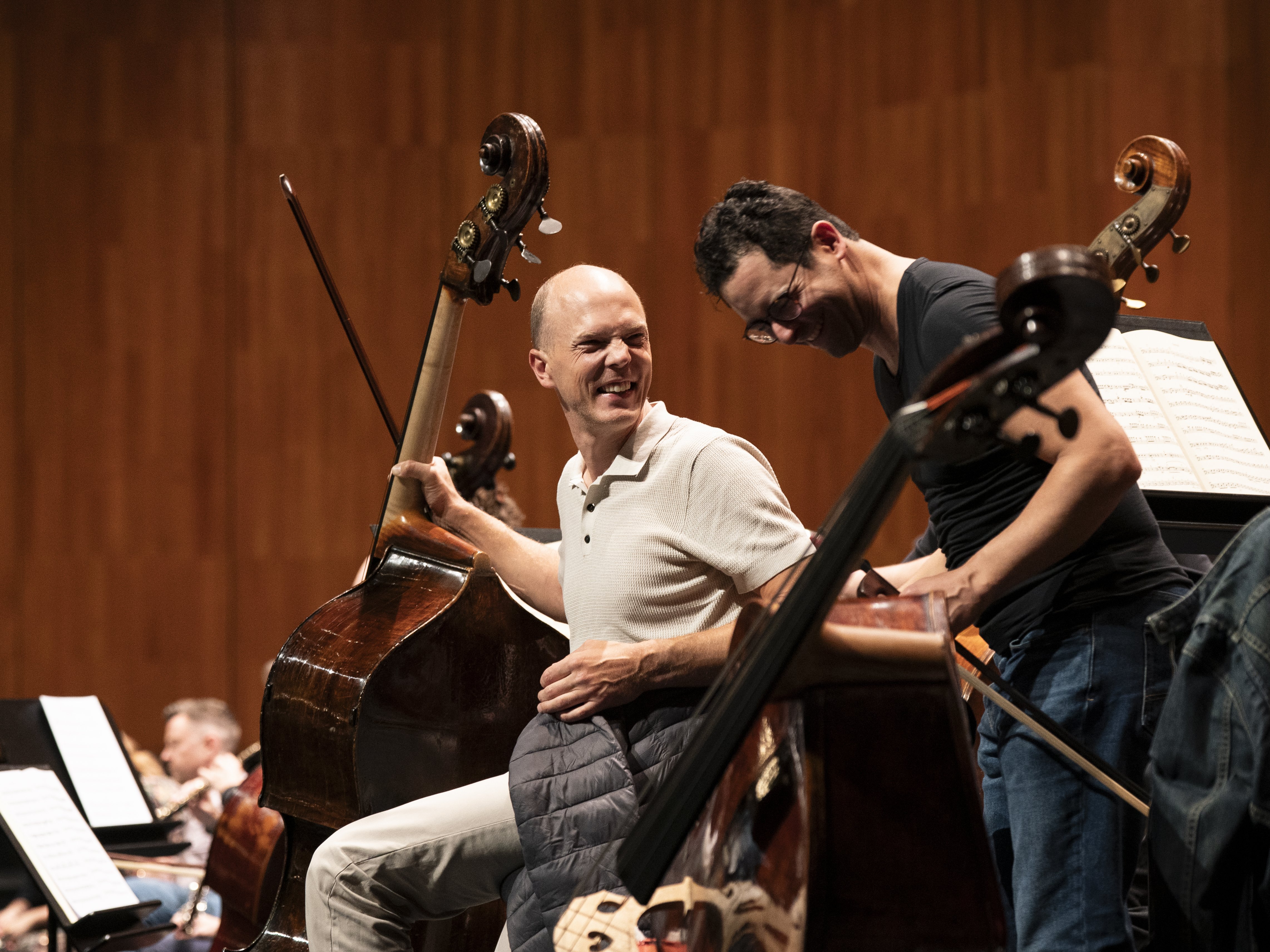 Zwei Musiker mit Kontrabässen lächeln und unterhalten sich während einer Probe auf der Bühne; im Hintergrund sind vor einer Holzwand weitere Orchestermitglieder und Notenständer zu sehen.