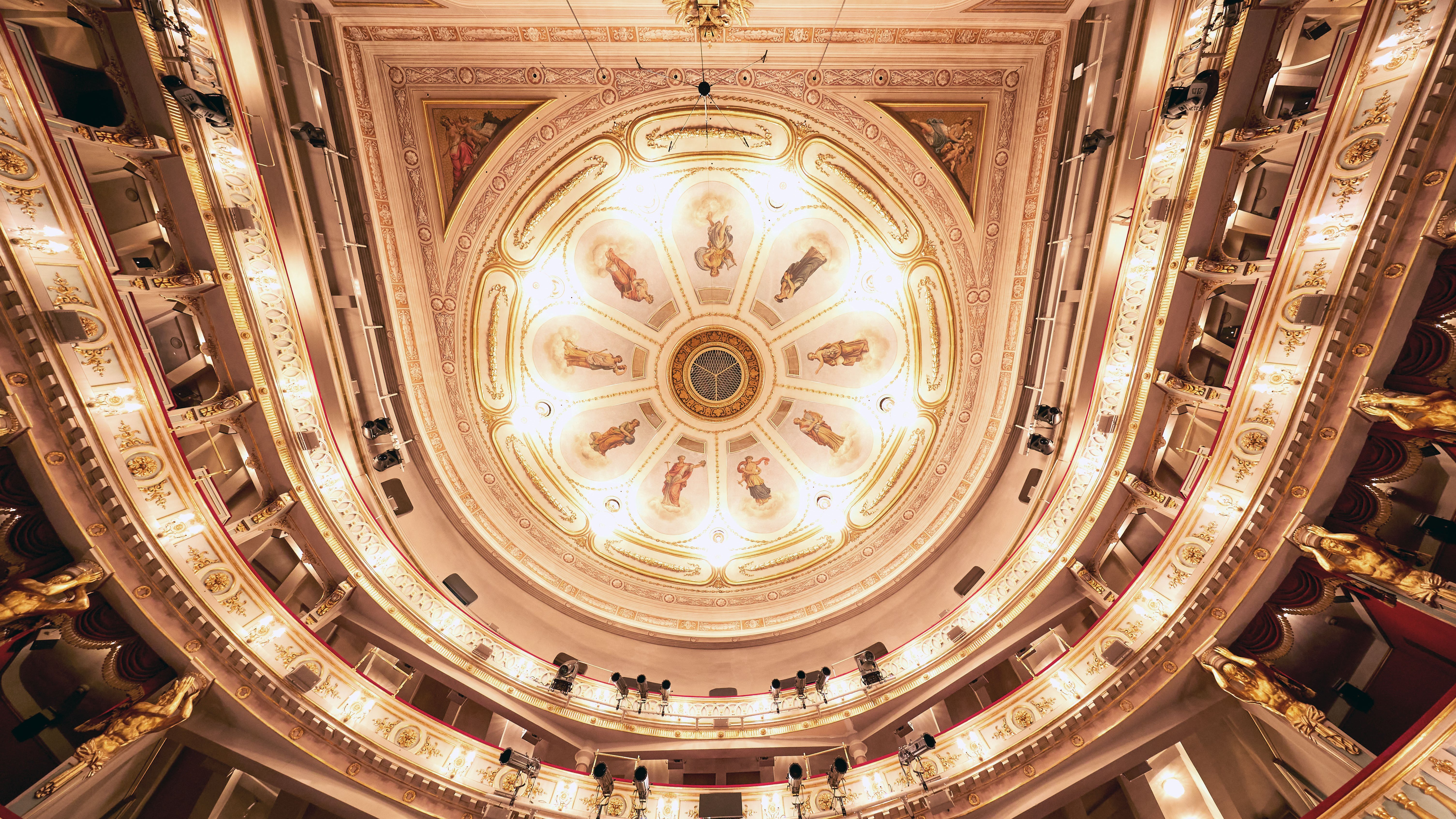 Verzierte, beleuchtete Decke des Theater an der Wien mit aufwendigen Dekorationen, bemalten Tafeln, goldenen Akzenten und um den Innenraum geschwungenen Balkonen, von unten betrachtet.