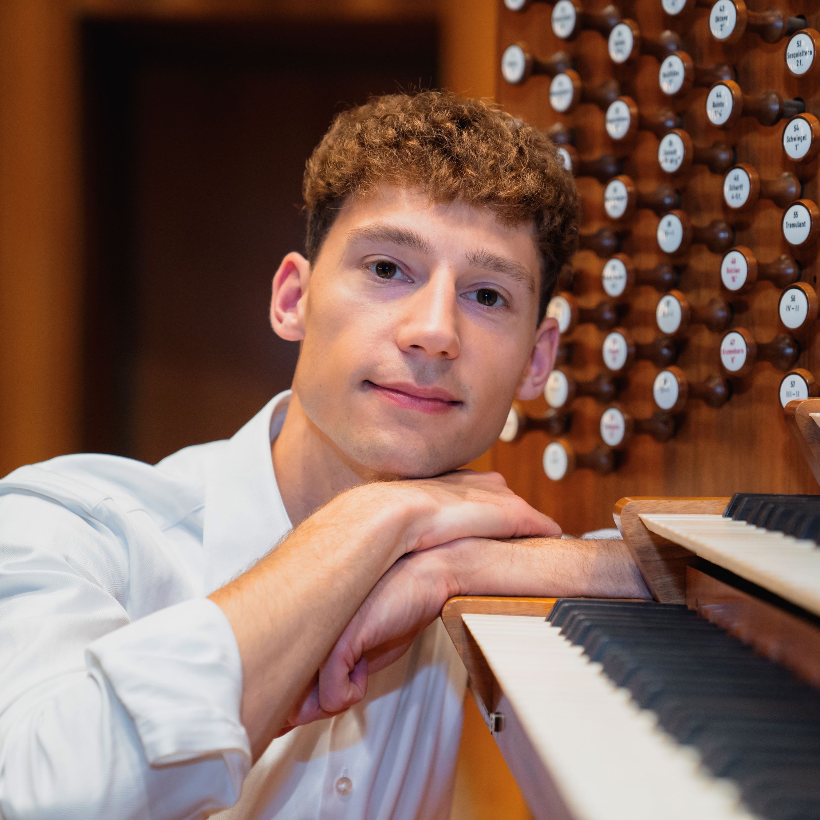Lukas Hasler im weißen Hemd stützt sein Kinn auf die Hände, die auf dem hölzernen Spieltisch einer Orgel ruhen. Neben ihm sind die Orgeltasten und Registerknöpfe sichtbar.