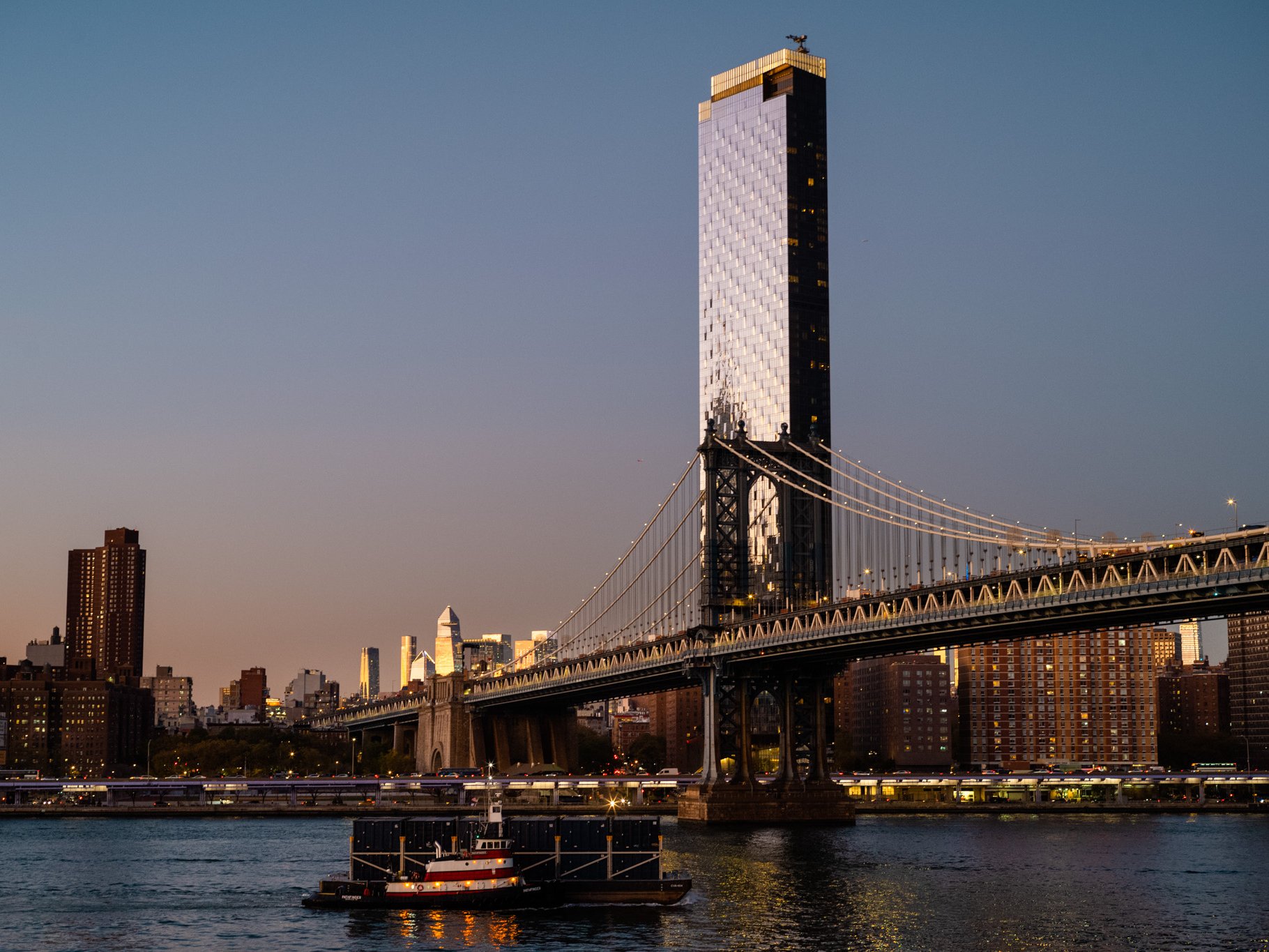 Ein Schlepper zieht in der Abenddämmerung auf dem East River vor der Manhattan Bridge und einem hohen Wolkenkratzer einen Lastkahn.