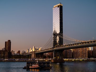The Manhattan Bridge spans the East River at twilight, with a sleek, reflective skyscraper rising behind it and a small tugboat navigating the water in the foreground.