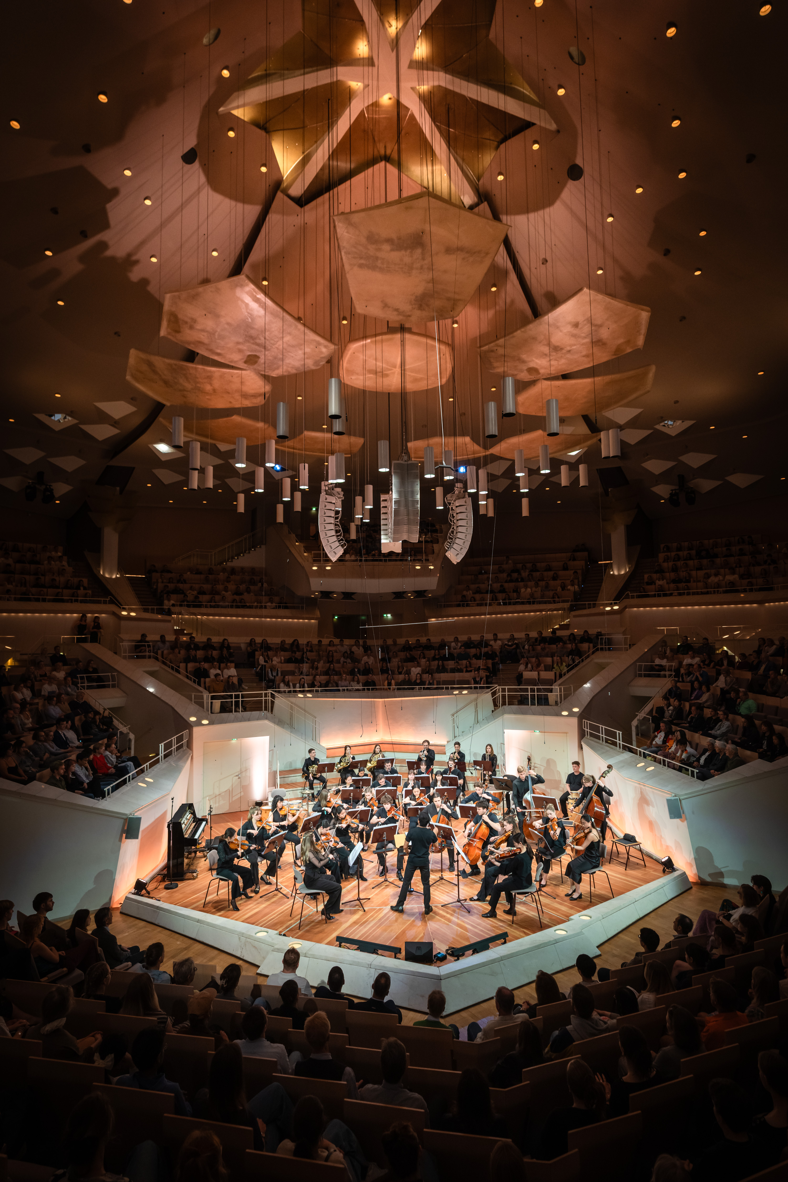 A symphony orchestra performs on stage in a modern concert hall filled with an audience. The hall features unique ceiling panels and dramatic lighting above the musicians.