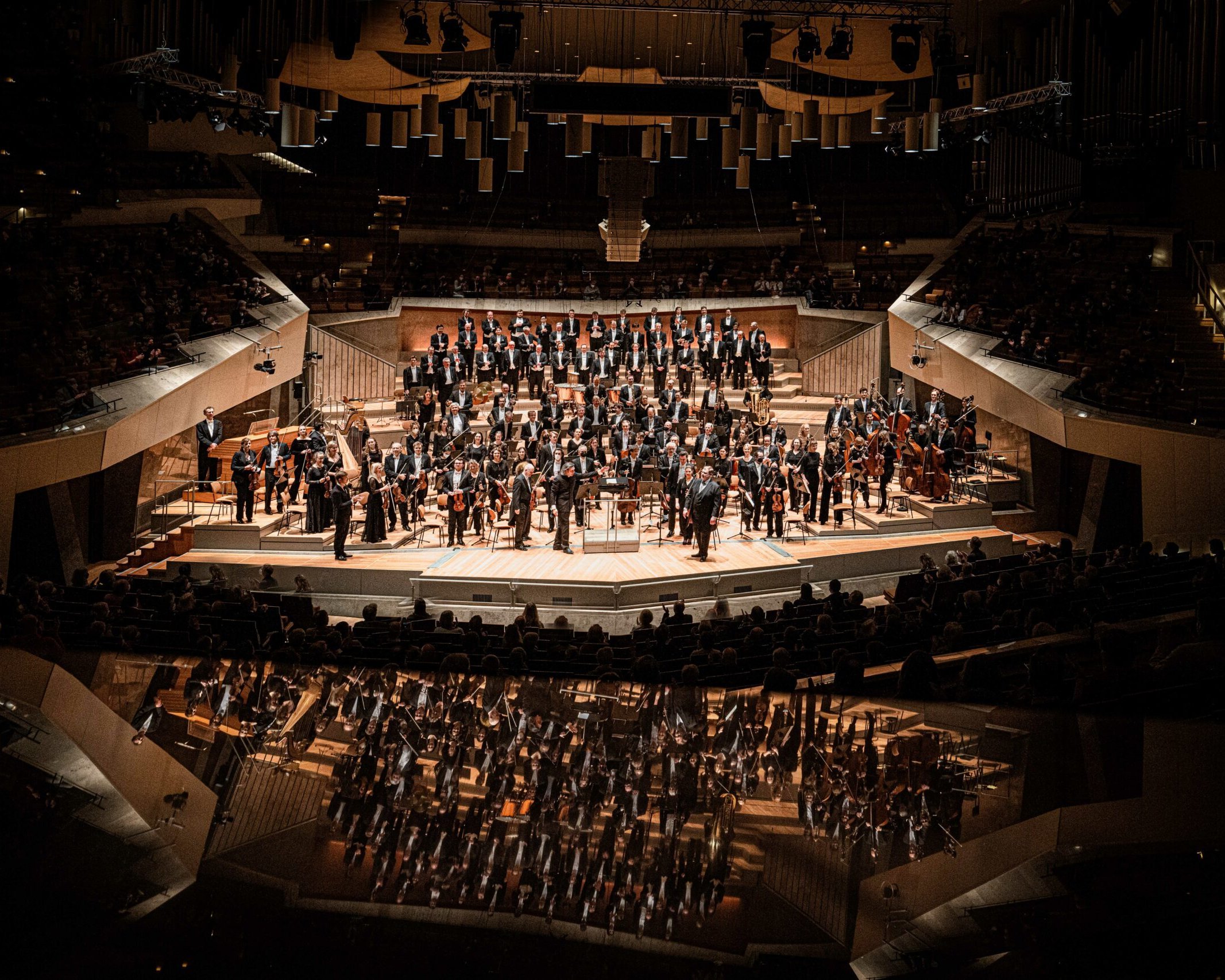 Ein großes Orchester spielt auf der Bühne eines großen Konzertsaals, wobei die Musiker in Sektionen sitzen und ihr Spiegelbild unten sichtbar ist, umgeben von einem Publikum bei gedämpfter Beleuchtung.