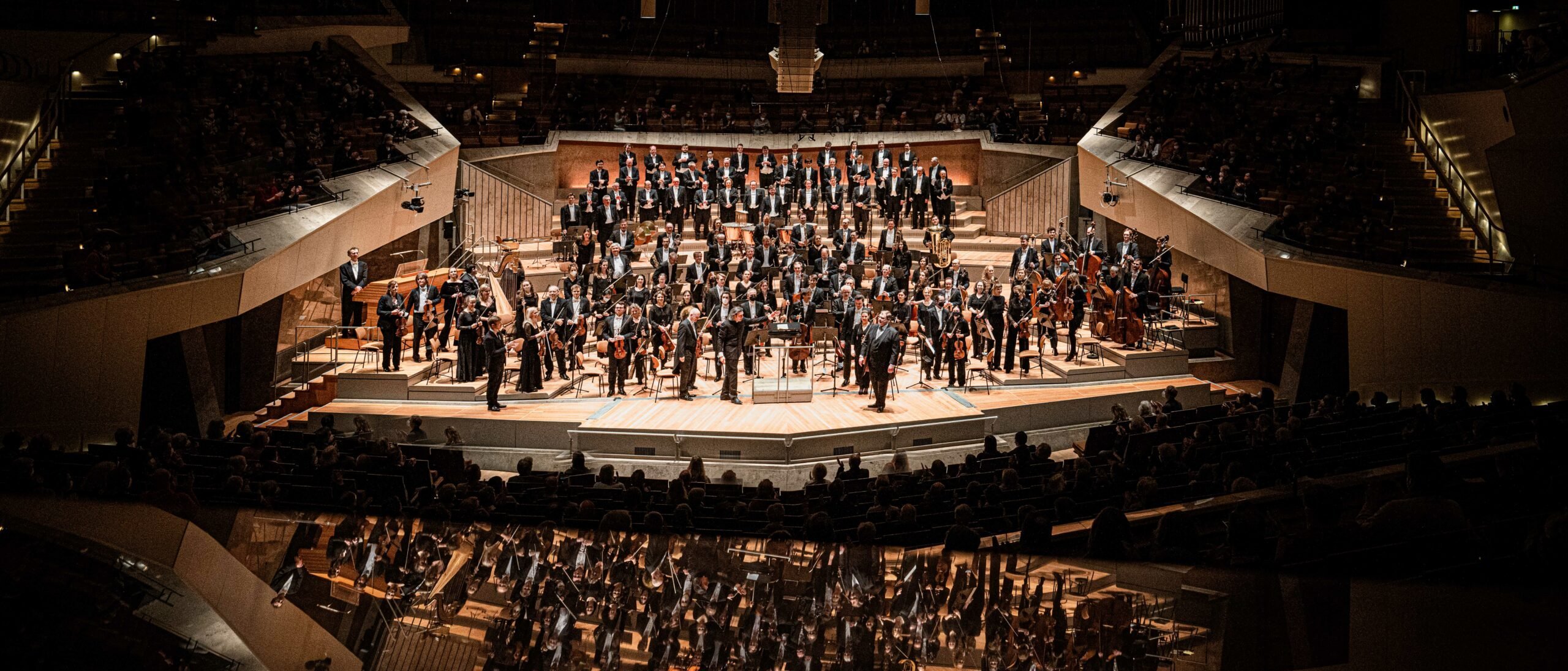 Ein großes Orchester spielt auf der Bühne eines großen Konzertsaals, wobei die Musiker in Sektionen sitzen und ihr Spiegelbild unten sichtbar ist, umgeben von einem Publikum bei gedämpfter Beleuchtung.