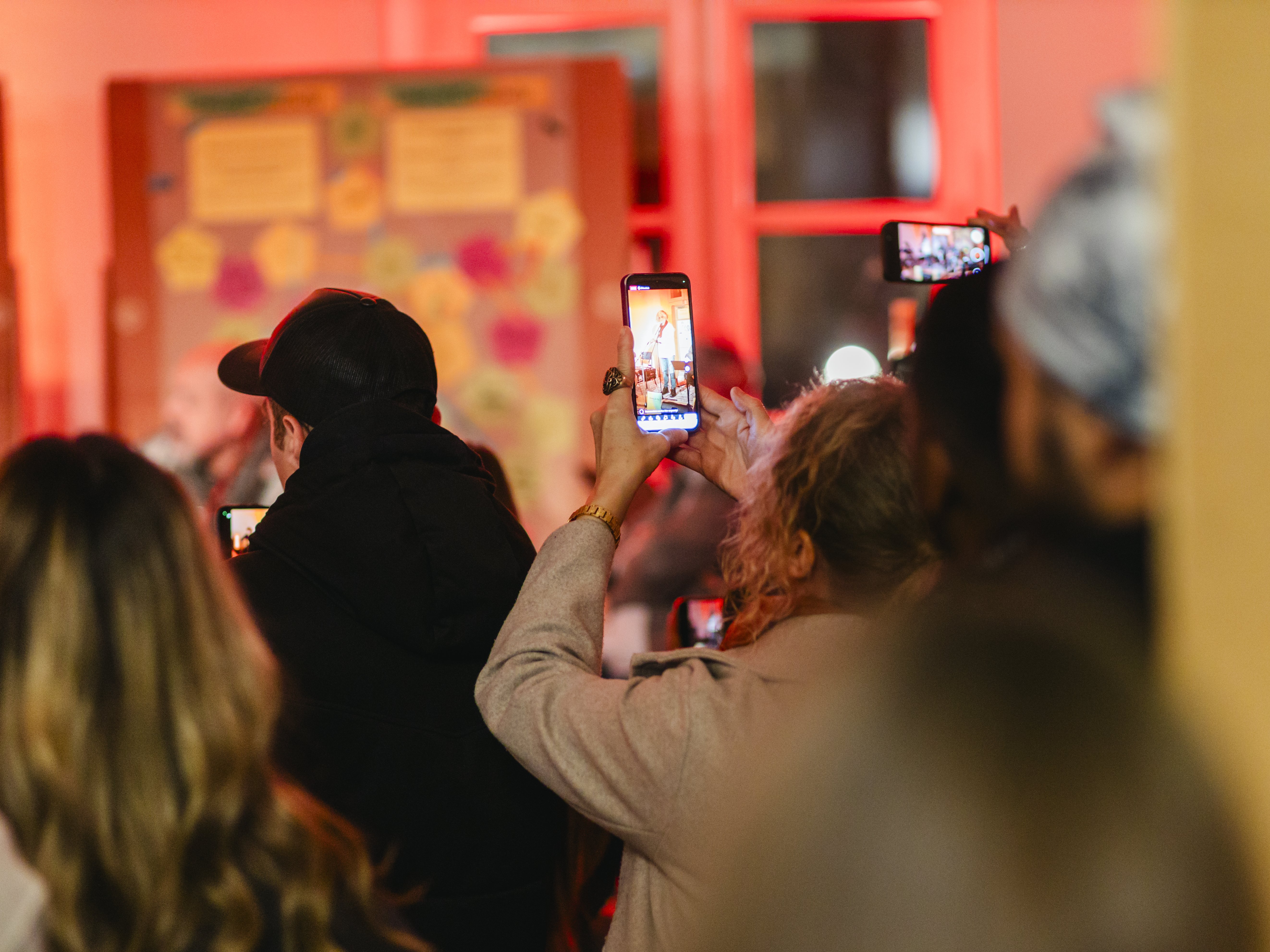 Eine Gruppe von Menschen steht in einem Innenraum und hält ihre Telefone hoch, um ein Ereignis aufzunehmen oder Fotos zu machen. Der Hintergrund ist mit roten Lichtern warm beleuchtet und mit Tafeln mit bunten Notizen geschmückt.