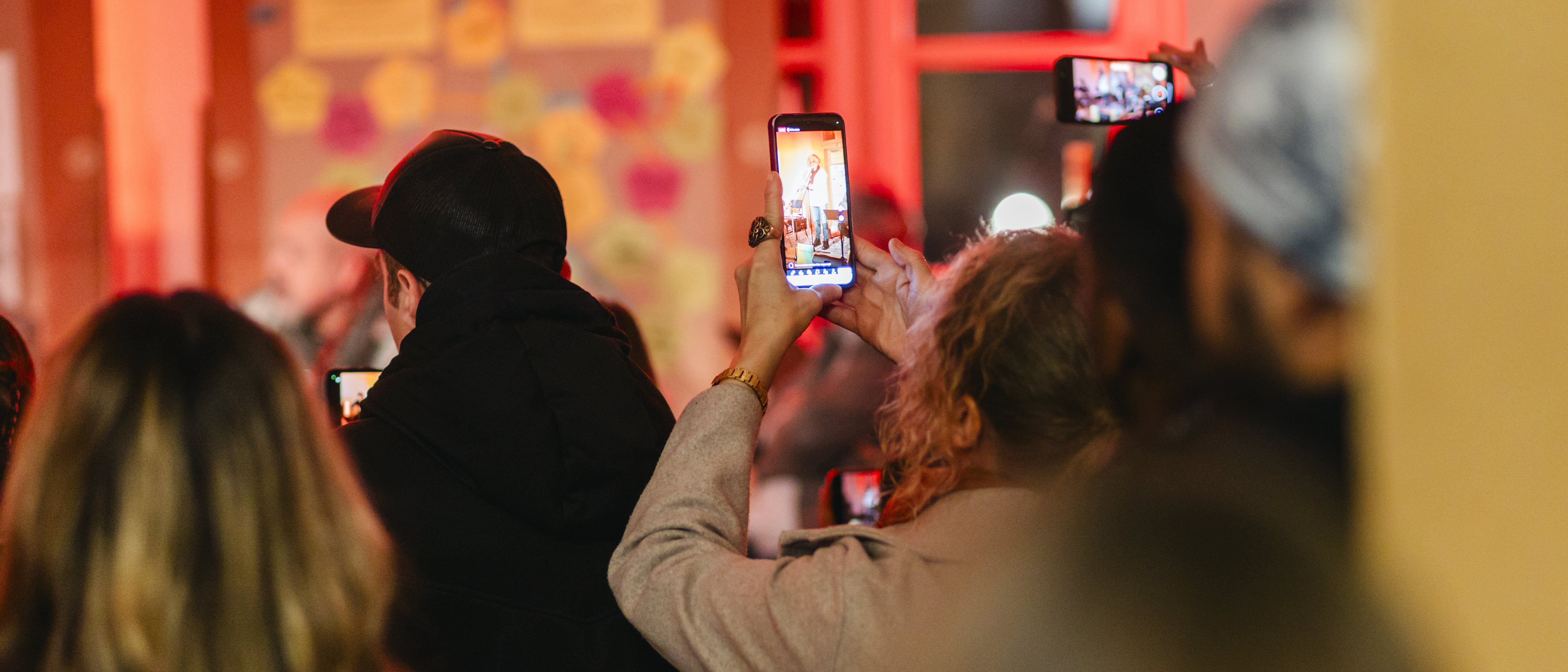 Eine Gruppe von Menschen steht in einem Innenraum und hält ihre Telefone hoch, um ein Ereignis aufzunehmen oder Fotos zu machen. Der Hintergrund ist mit roten Lichtern warm beleuchtet und mit Tafeln mit bunten Notizen geschmückt.