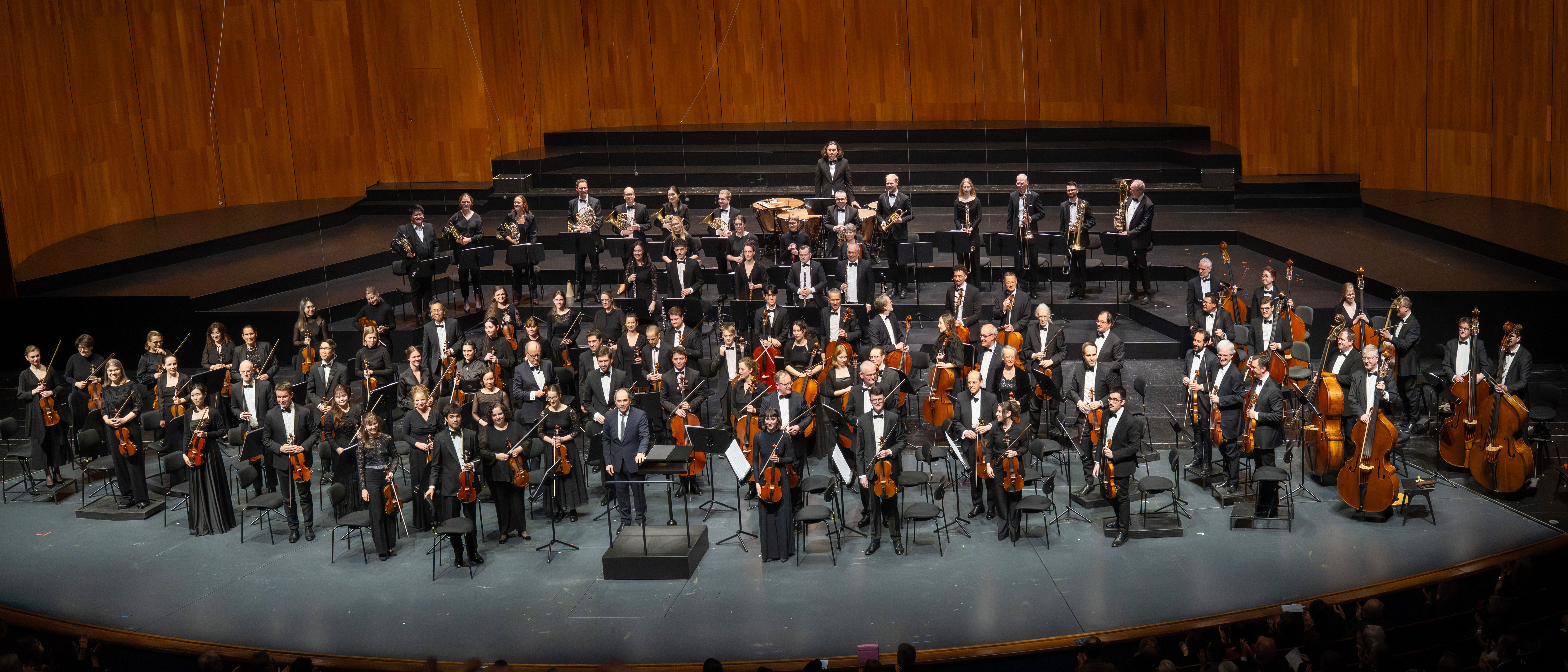 Ein Orchester steht mit seinen Instrumenten auf der Bühne und blickt auf das Publikum in einem modernen Konzertsaal mit einem Holzhintergrund. Die Musiker tragen formelle schwarze Kleidung und im Publikum sitzen Menschen.
