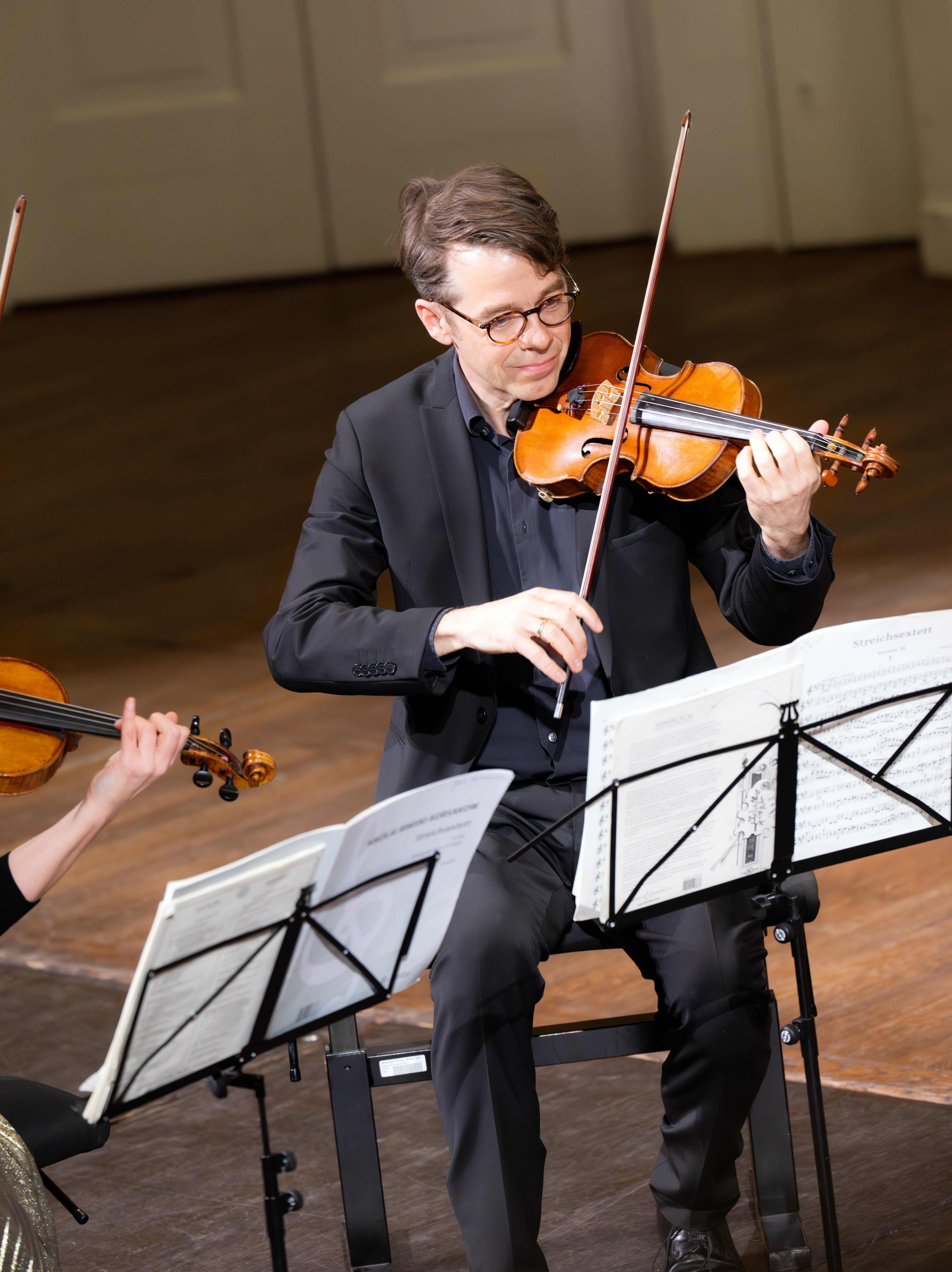 Ein Mann in einem schwarzen Anzug und mit Brille spielt auf der Bühne Geige, umgeben von Notenständern mit Notenblättern, während einer klassischen Musikaufführung.