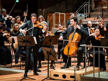 Noah Bendix-Balgley und Bruno Delepelaire treten mit einem Orchester in einem Konzertsaal auf - einer spielt Geige, einer Cello -, beide schwarz gekleidet, lächelnd, mit Notenständern vor ihnen und Streichern im Hintergrund.