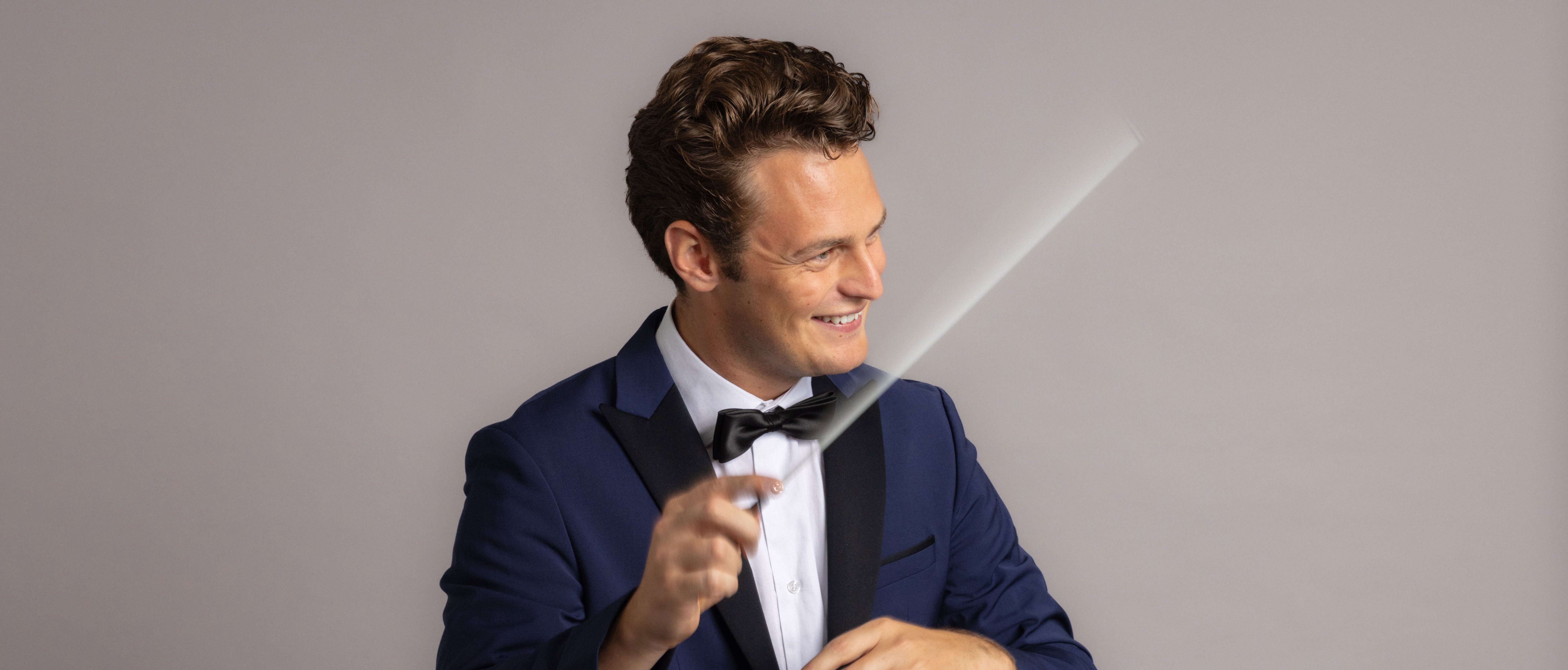 Ein lächelnder Mann im Smoking, der einen Dirigentenstab in der Hand hält.