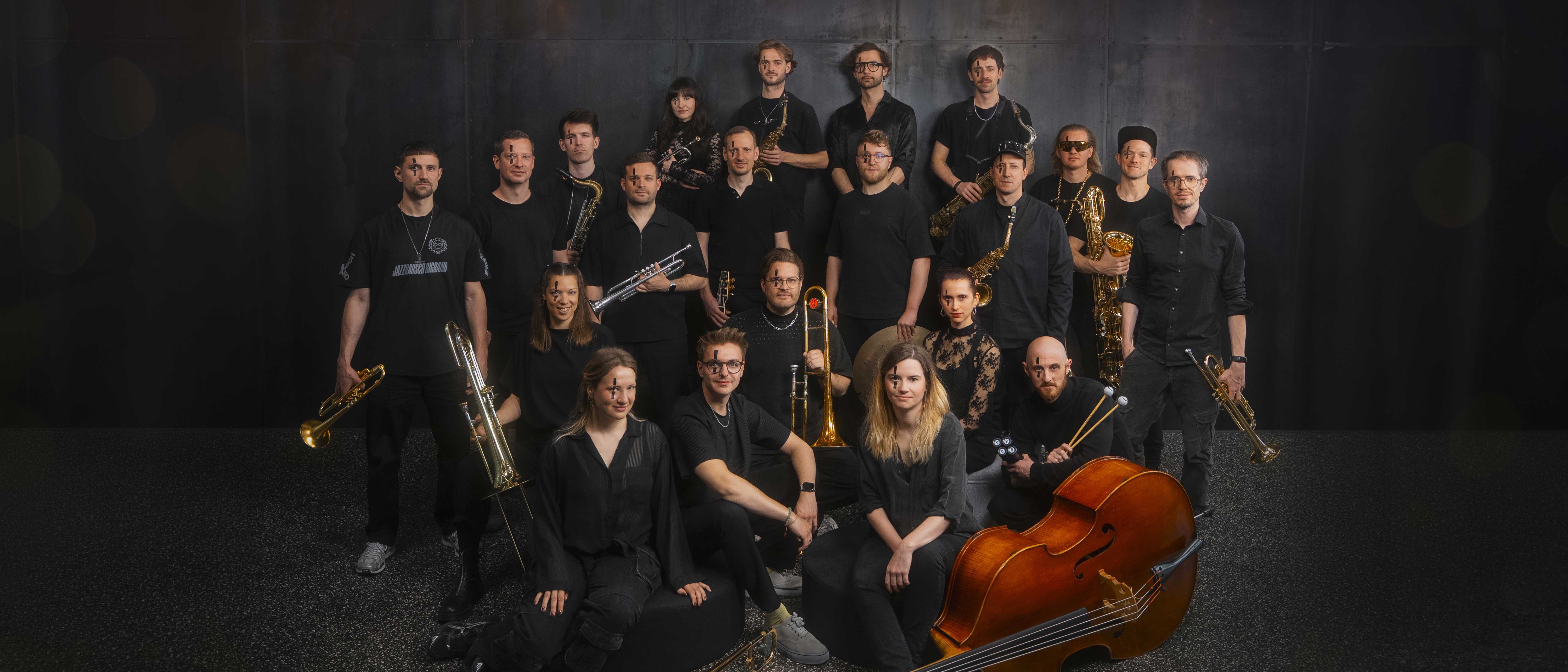 Eine Gruppe von 20 Musikern in schwarzer Kleidung posiert mit Blechblas-, Holzblas- und Streichinstrumenten vor einem dunklen Studiohintergrund.