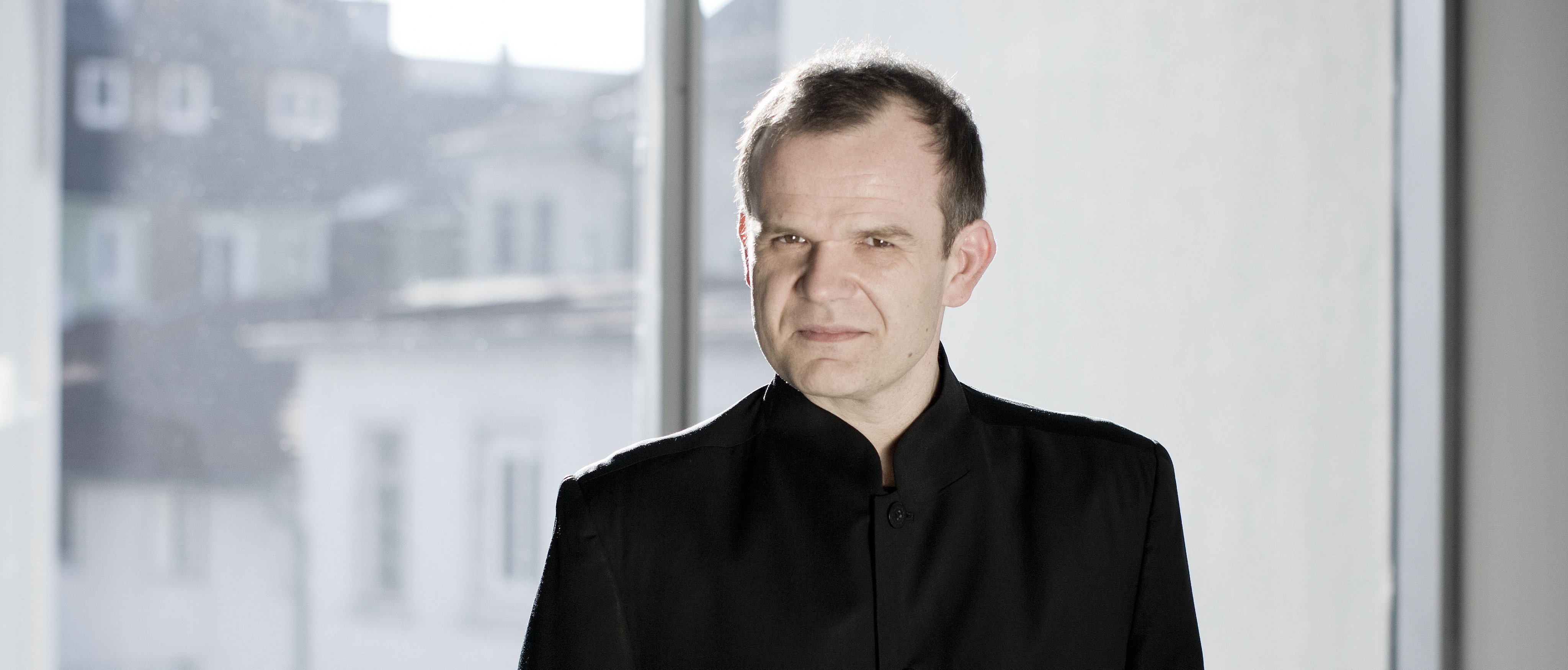Ein Mann mit kurzen Haaren steht vor einem großen Fenster. Er trägt einen schwarzen Anzug, hat die Hände in den Hosentaschen und schaut in die Kamera.