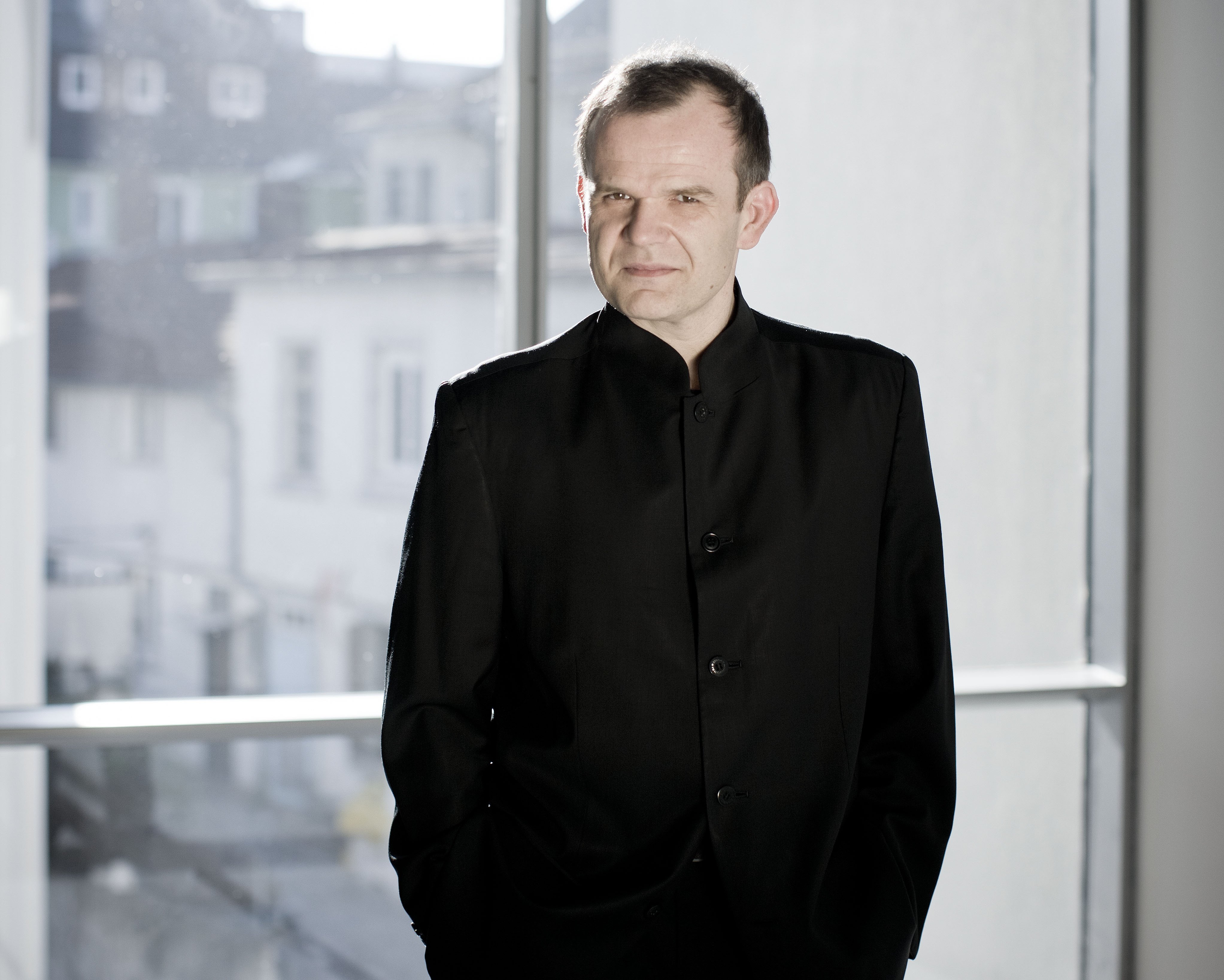 Ein Mann mit kurzen Haaren steht vor einem großen Fenster. Er trägt einen schwarzen Anzug, hat die Hände in den Hosentaschen und schaut in die Kamera.