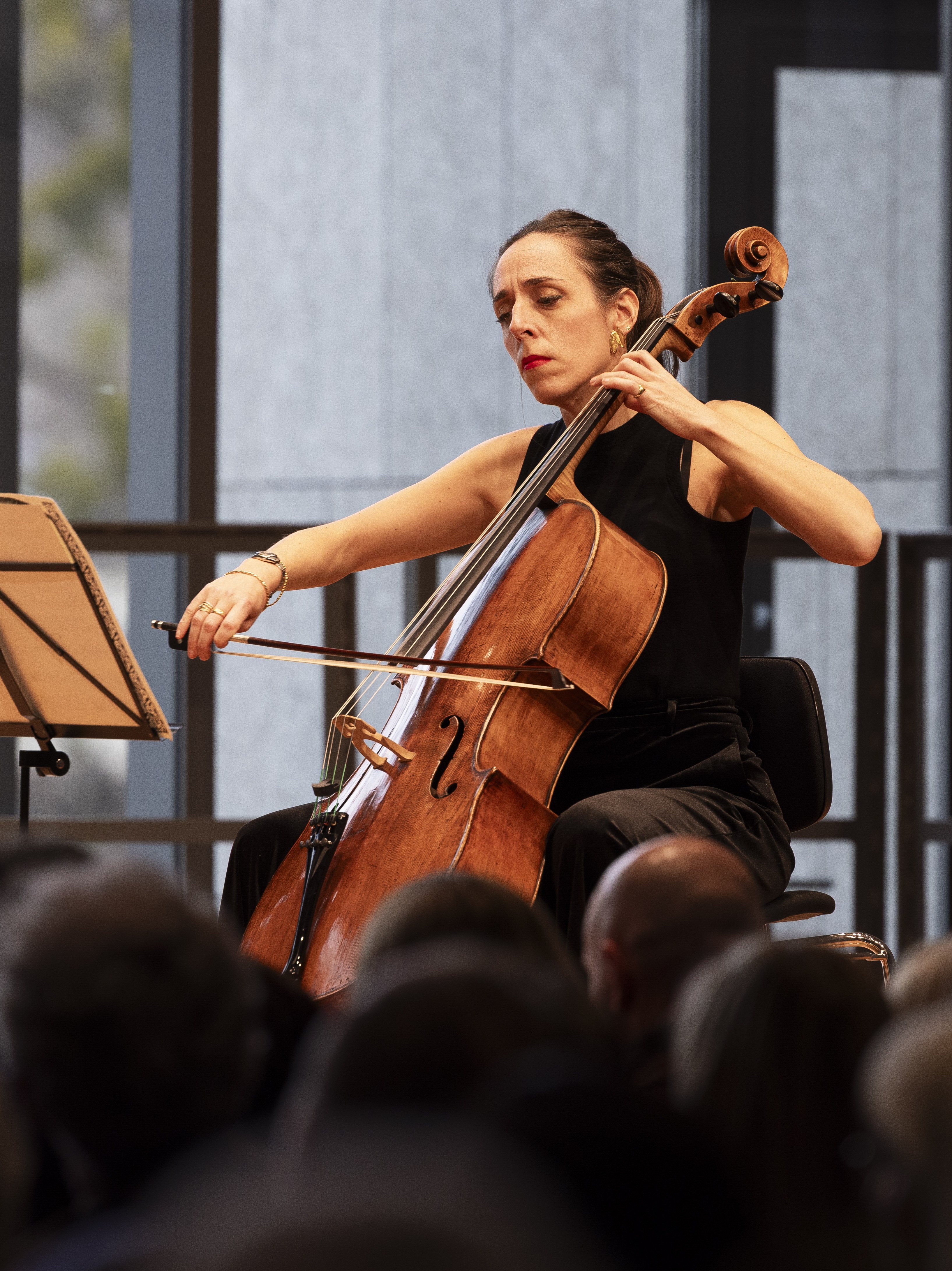 Eine Frau in einem schwarzen ärmellosen Oberteil spielt auf der Bühne konzentriert Cello. Sie sitzt vor einem offenen Notenpult, mit einem Publikum und großen Fenstern im Hintergrund.