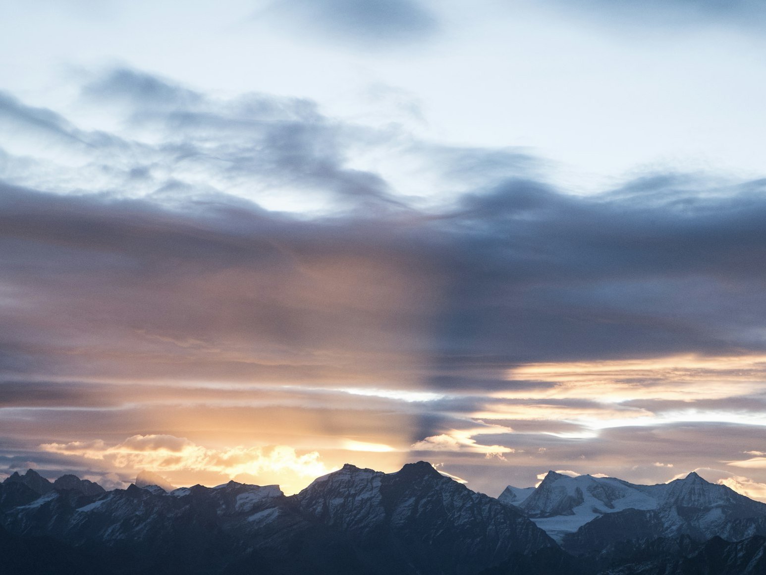 Sunset over snow-capped mountains, with rays of sunlight streaming through dramatic clouds, casting shadows and warm light across the sky.