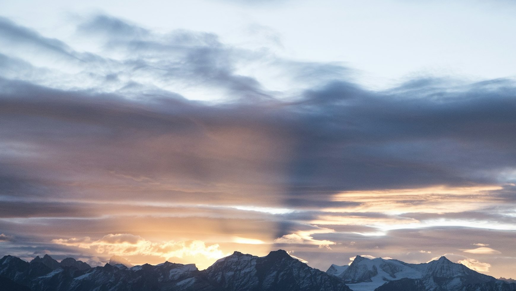 Sunset over snow-capped mountains, with rays of sunlight streaming through dramatic clouds, casting shadows and warm light across the sky.