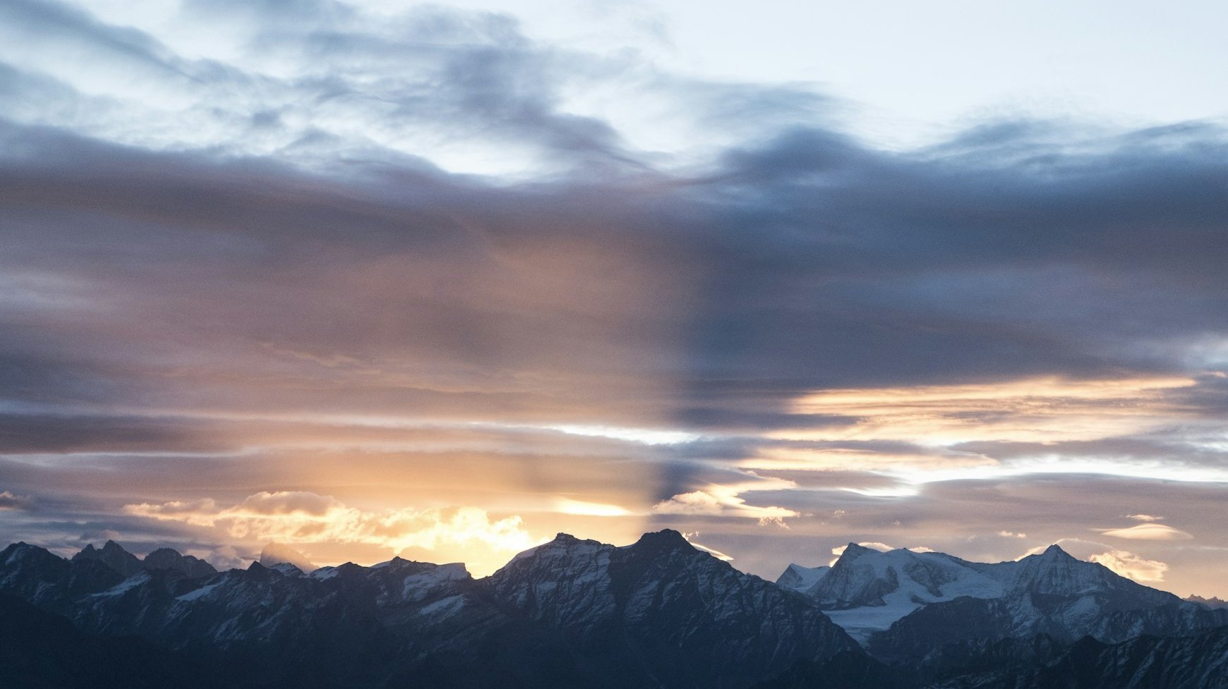 Verschneite Berggipfel unter einem dramatischen Himmel, an dem bei Sonnenaufgang oder Sonnenuntergang Sonnenstrahlen und Schatten durch bunte Wolken brechen.