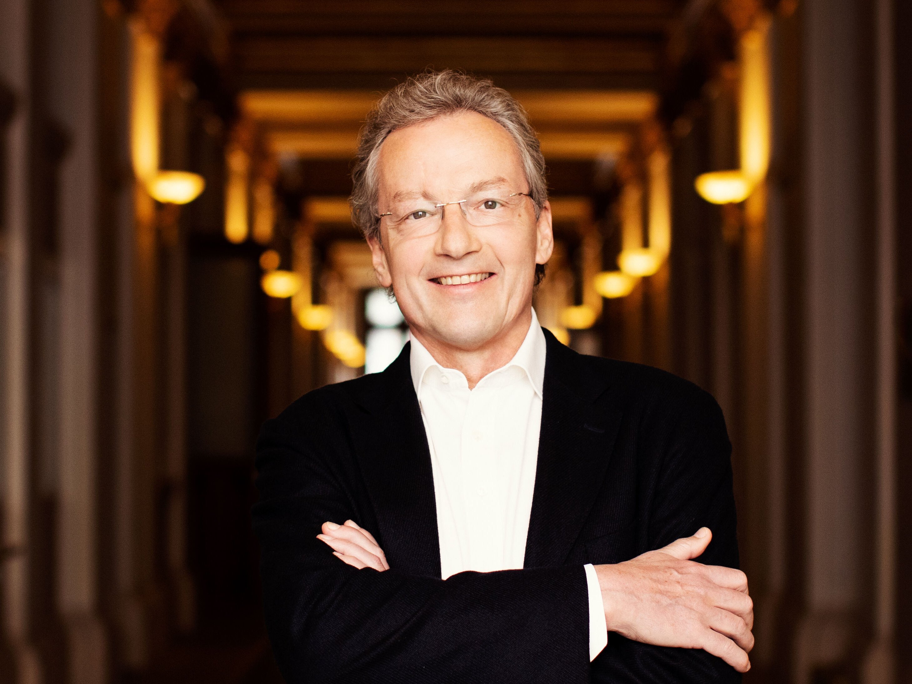 Franz Welser-Möst mit Brille und grauem Haar lächelt. Er trägt ein weißes Hemd und ein dunkles Jackett. Mit verschränkten Armen steht er in einem sanft beleuchteten Flur mit dekorativen Säulen.