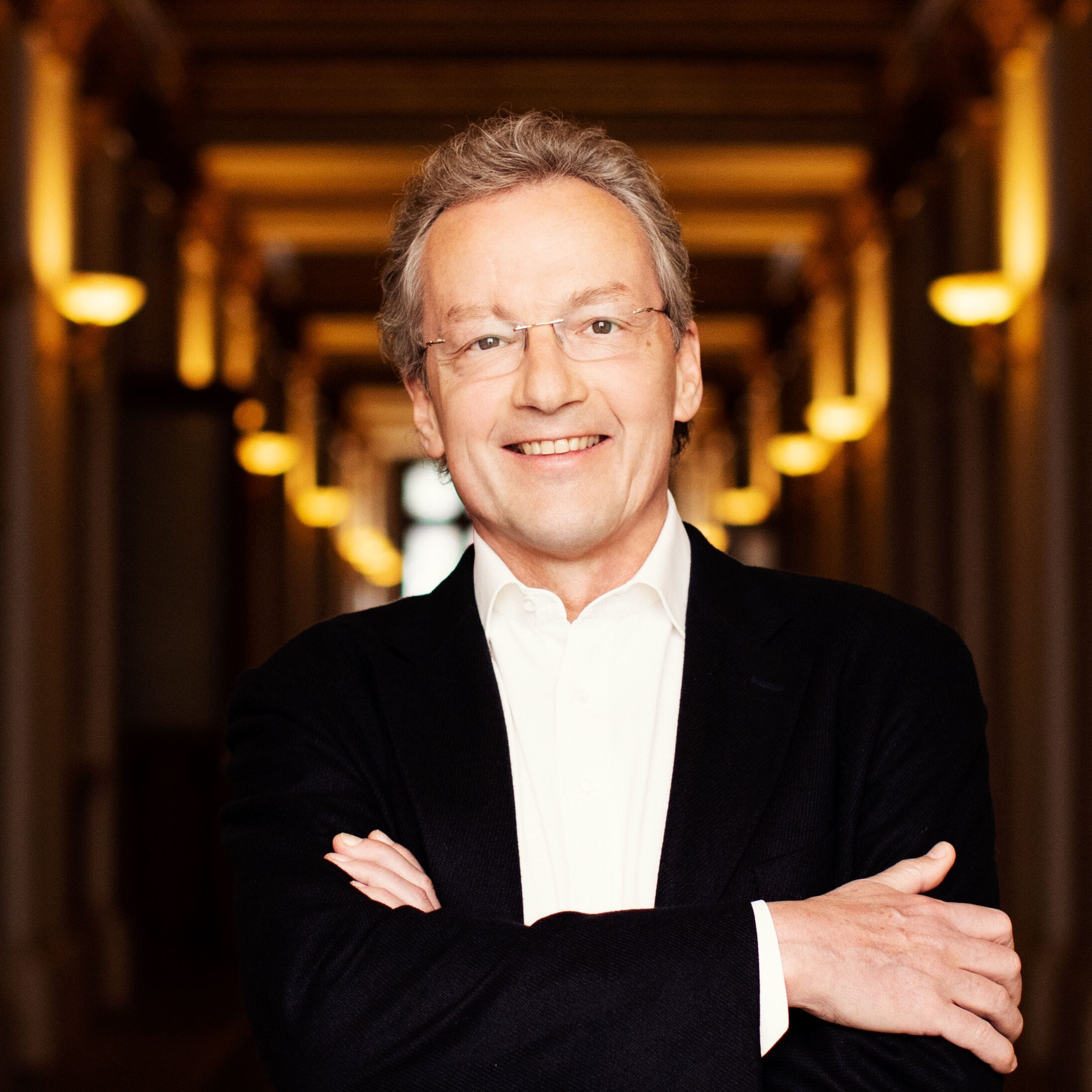 Franz Welser-Möst mit Brille und grauem Haar lächelt. Er trägt ein weißes Hemd und ein dunkles Jackett. Mit verschränkten Armen steht er in einem sanft beleuchteten Flur mit dekorativen Säulen.