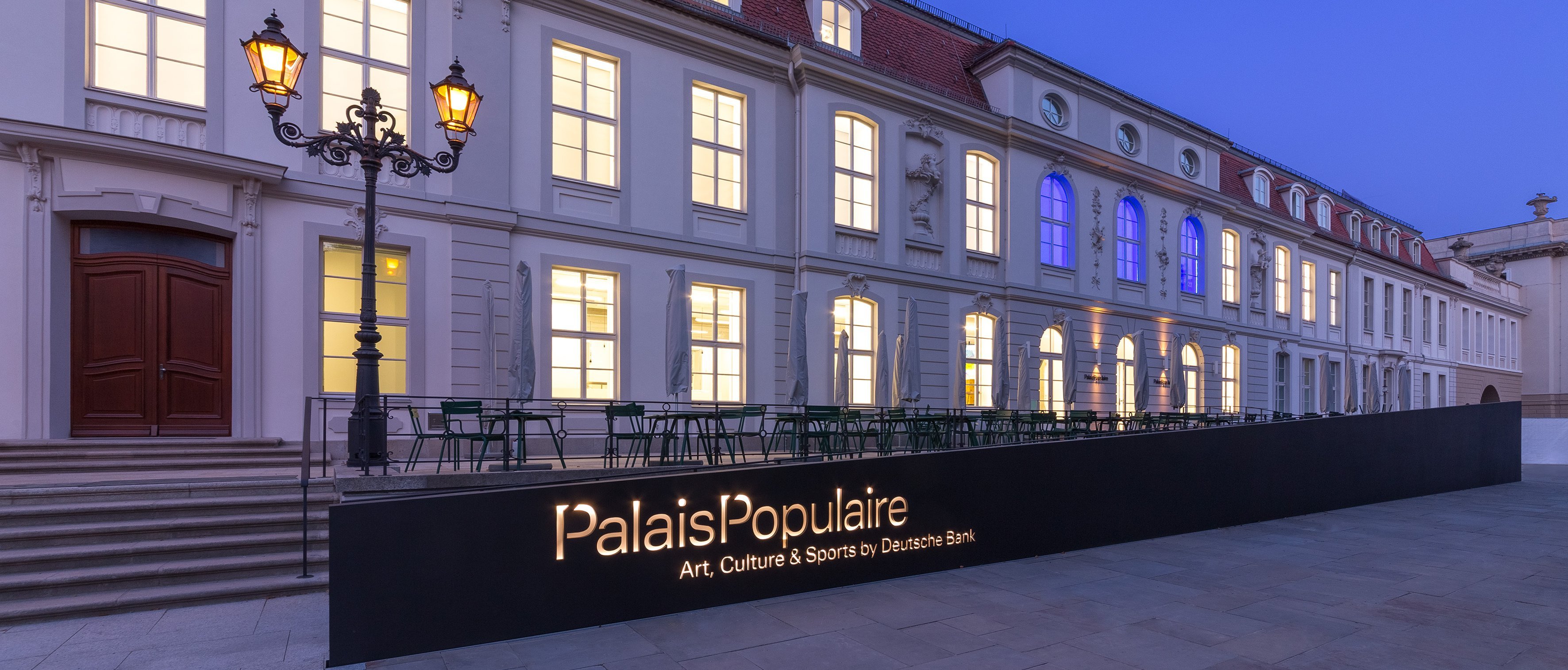 Ein modernes, gut beleuchtetes Gebäude in der Abenddämmerung mit großen Fenstern, einem Schild mit der Aufschrift PalaisPopulaire Art, Culture & Sports by Deutsche Bank und Sitzgelegenheiten im Freien neben einer alten Straßenlaterne.
