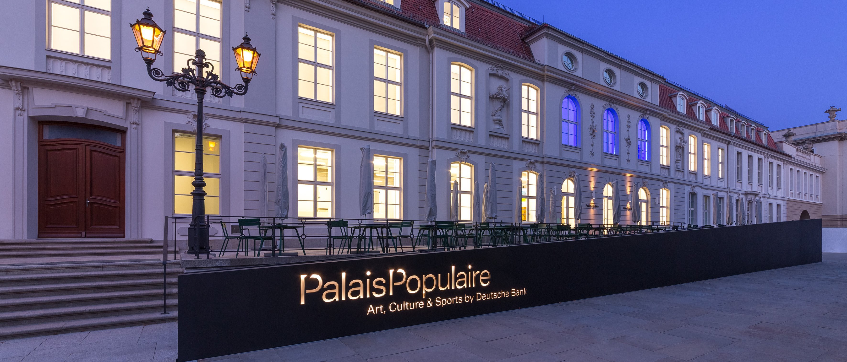 A modern illuminated sign for Palais Populaire stands in front of a grand historic building with large windows glowing at dusk under a clear blue sky.