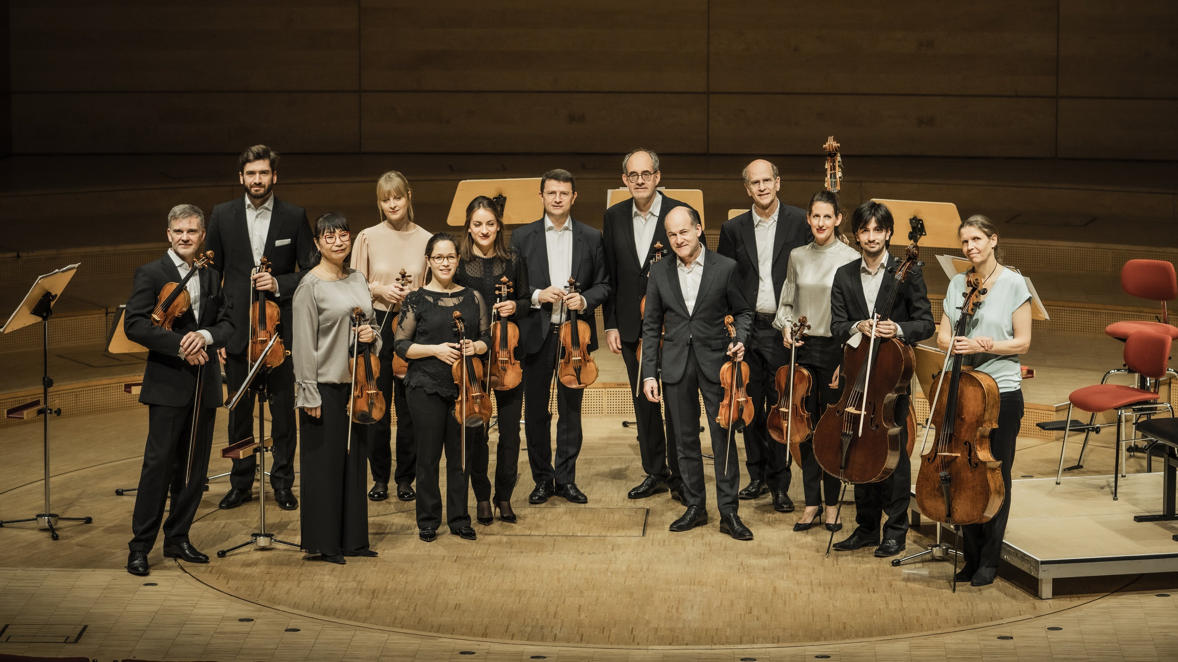 A group of thirteen musicians holding string instruments stands on a wooden concert stage, posing and smiling for a photo. Several empty chairs and music stands are visible in the background.