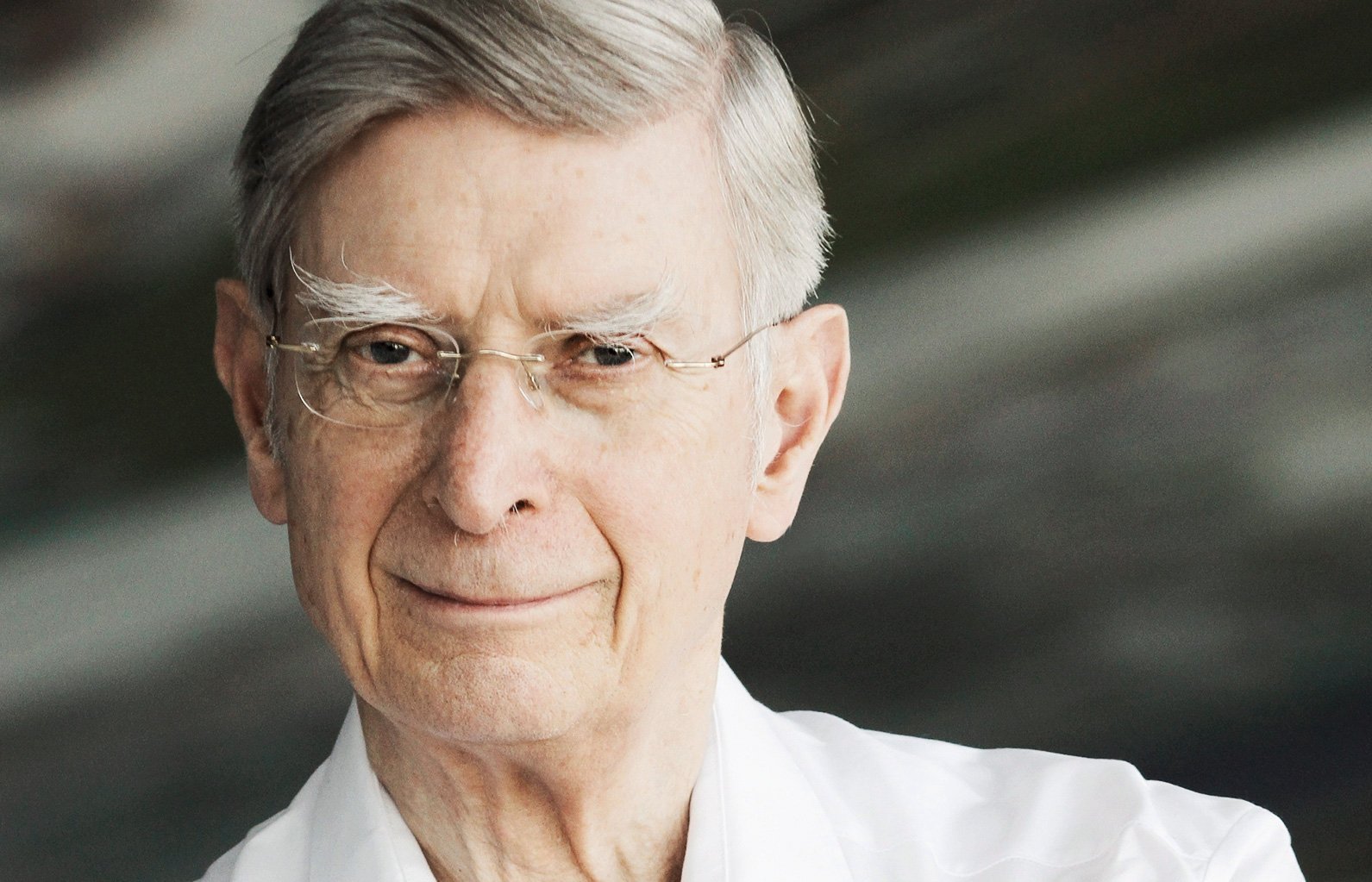 Herbert Blomstedt mit grauem Haar und Brille, trägt ein weißes Hemd und lächelt sanft, während er in die Kamera blickt. Der Hintergrund ist unscharf.