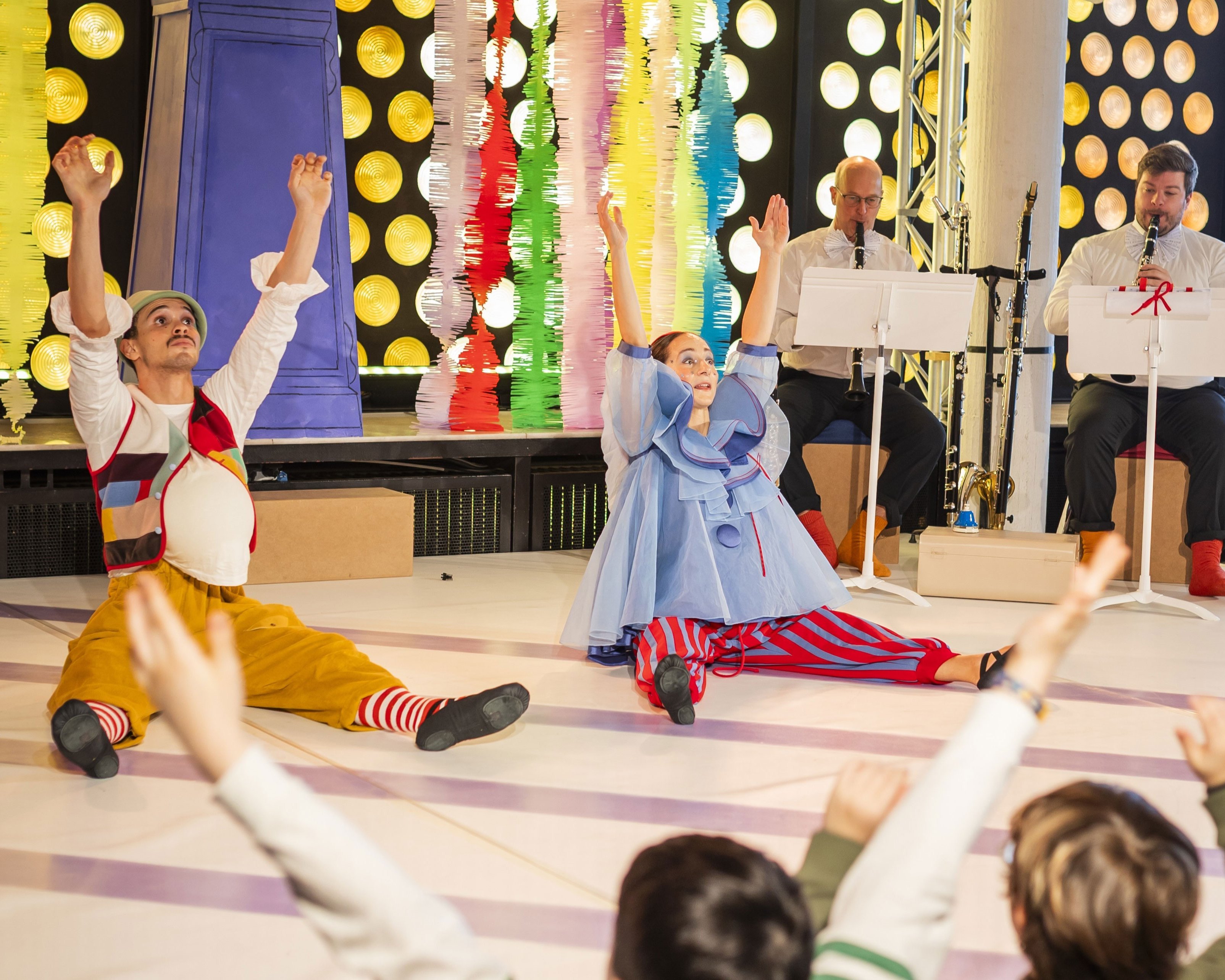 Zwei als Clowns verkleidete Darsteller sitzen mit erhobenen Armen auf dem Boden und unterhalten Kinder vor einer farbenfrohen, festlichen Kulisse. Im Hintergrund spielen Musiker, und die Kinder ahmen die Aktionen der Darsteller nach.