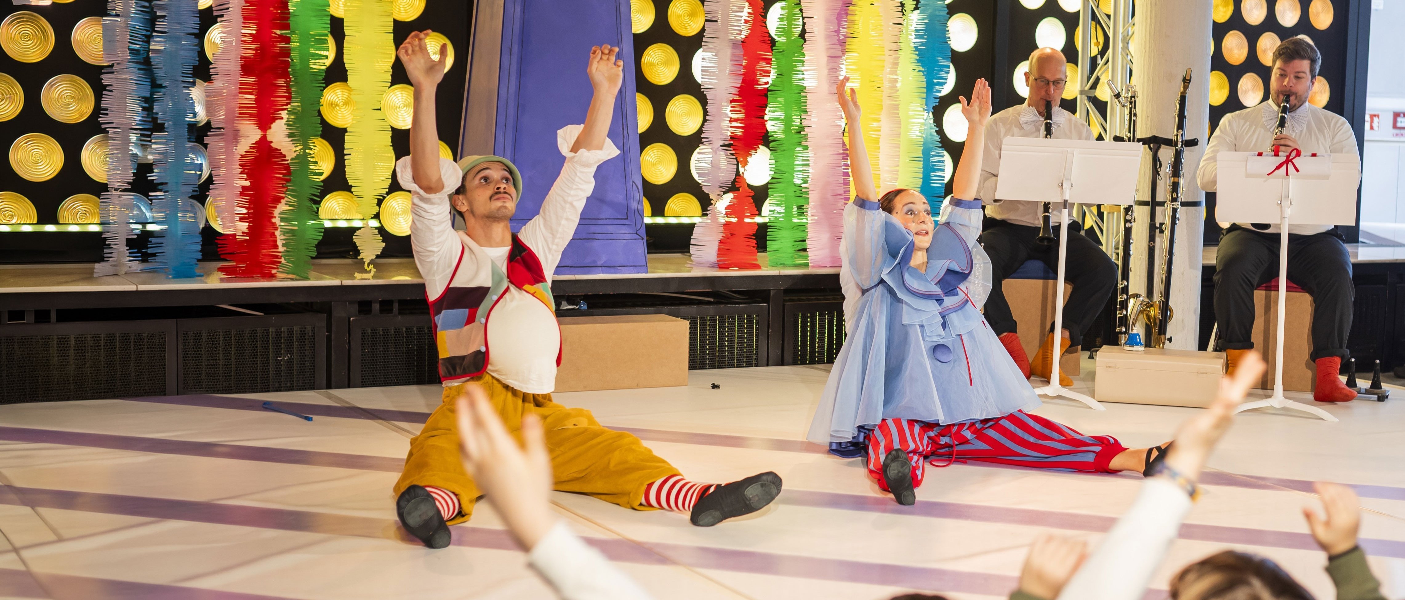 Zwei als Clowns verkleidete Darsteller sitzen mit erhobenen Armen auf dem Boden und unterhalten Kinder vor einer farbenfrohen, festlichen Kulisse. Im Hintergrund spielen Musiker, und die Kinder ahmen die Aktionen der Darsteller nach.
