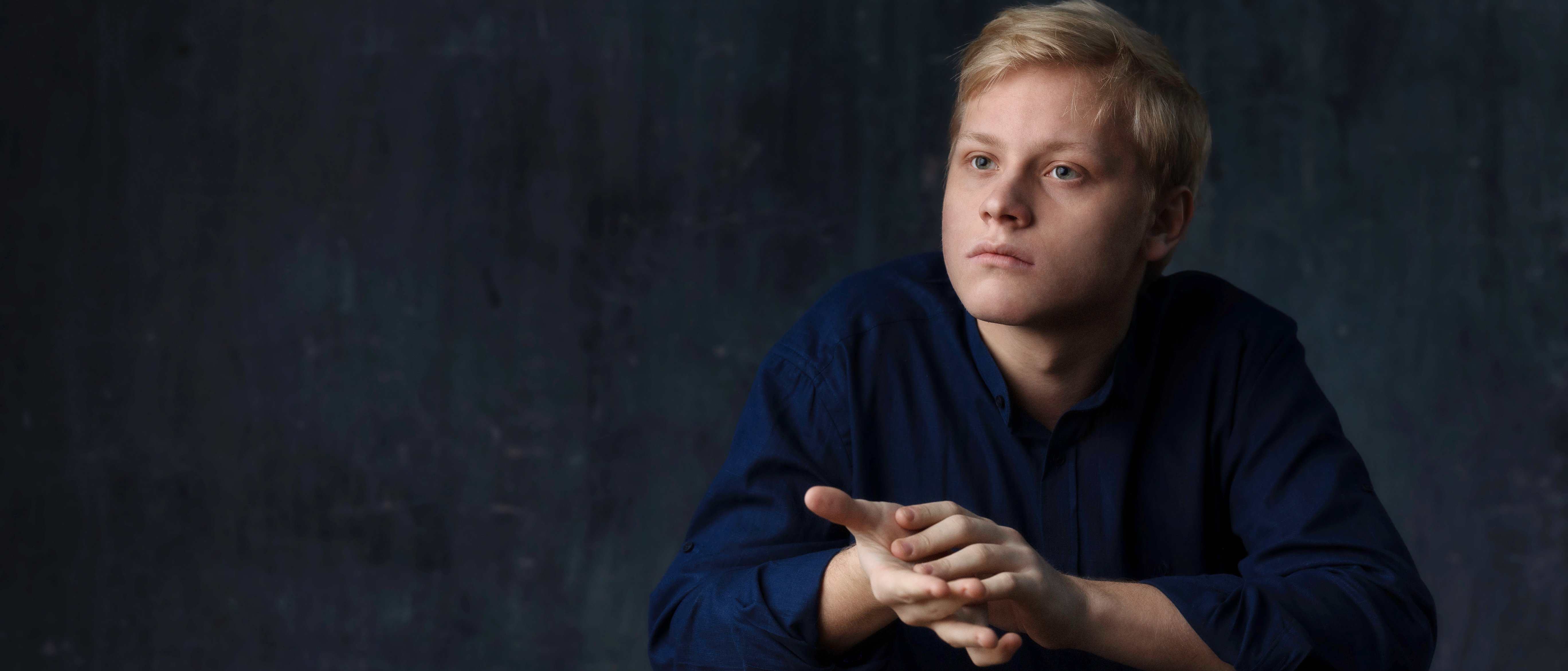 Alexander Malofeev mit blondem Haar und dunkelblauem Hemd sitzt mit gefalteten Händen an einem Holztisch und blickt nachdenklich zur Seite vor einem dunklen, strukturierten Hintergrund.