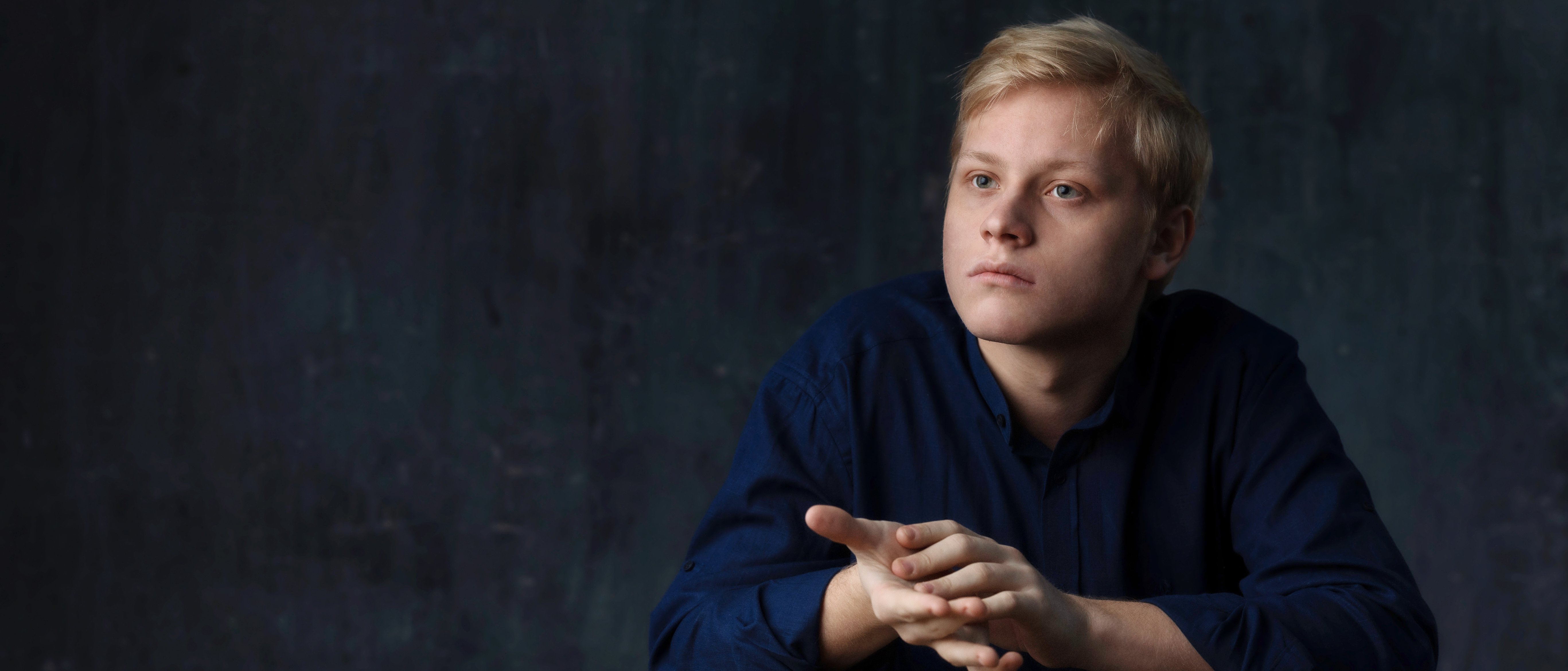 Alexander Malofeev with short blond hair, wearing a dark blue shirt, sits at a round wooden table against a dark, textured background, looking thoughtfully into the distance.