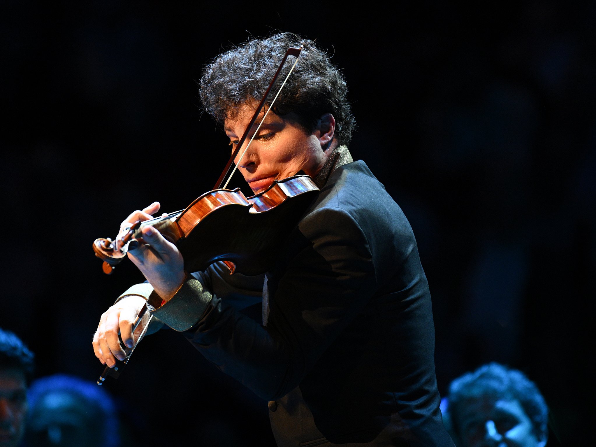 A violinist in a dark suit plays passionately on stage, illuminated by a spotlight, with a focused expression. Blurred audience members are visible in the background.