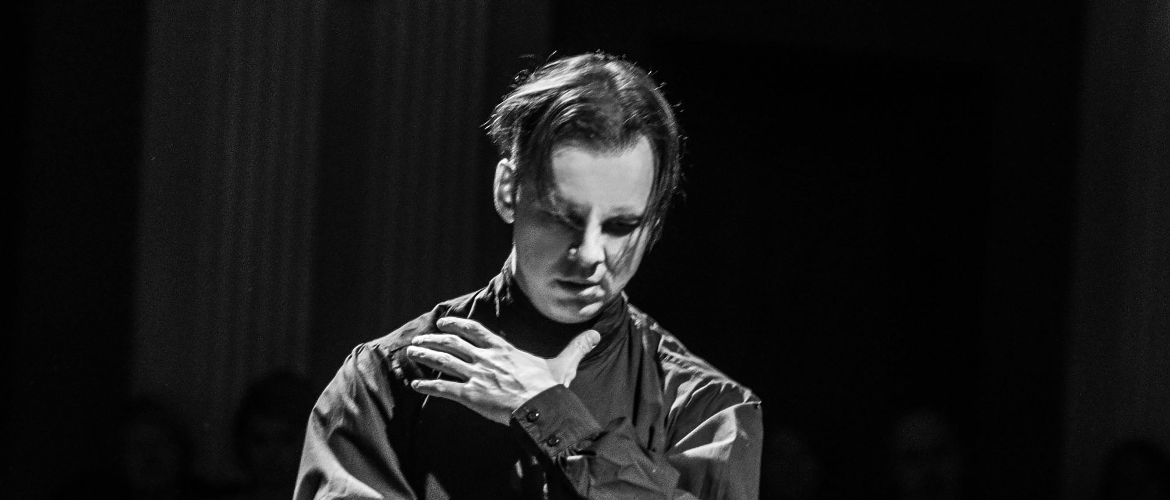 A person conducting music with a serious expression, one hand on their chest and the other raised. The image is in black and white, and the background is dimly lit with faint audience figures.