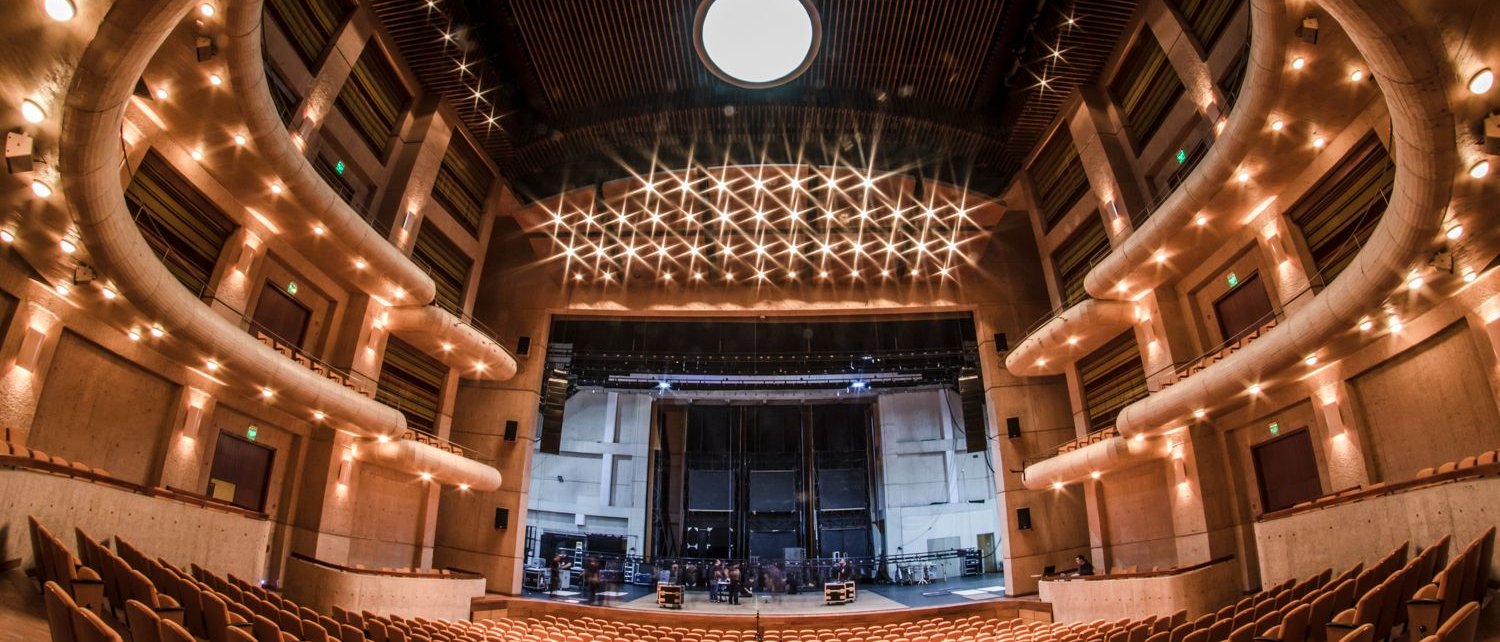 A wide-angle view of an empty modern theater auditorium with rows of tan seats, curved balconies, and a large stage under bright ceiling lights.
